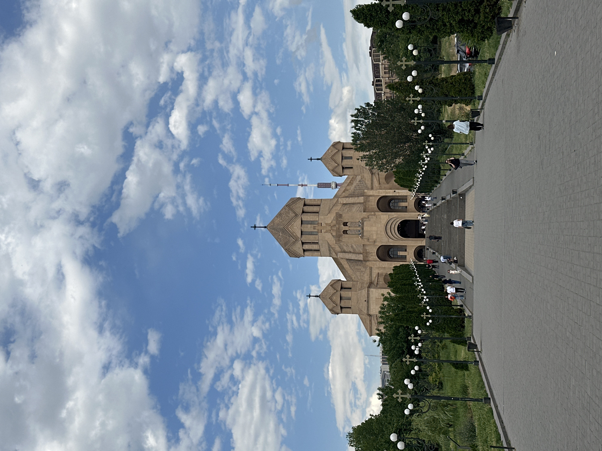 The image, when correctly oriented, depicts a grand outdoor scene dominated by a large, light-brown stone church under a bright blue sky filled with scattered white cumulus clouds. The church features multiple pointed domes and towers, each topped with a cross, characteristic of Armenian ecclesiastical architecture. Arched entrances are visible on the facade.
A wide, paved pathway made of grey brick-like tiles stretches towards the church, creating a sense of perspective. On either side of this pathway are neatly maintained green lawns and rows of trees. Numerous black lampposts line the edges of the pathway, fitted with spherical white lights and decorative elements, some of which are shaped like crosses.
Several individuals are visible on the pathway, appearing small against the backdrop of the large church and extensive grounds. They are casually dressed and seem to be walking or strolling, suggesting general foot traffic or visitors to the site. Behind the church, a tall, slender structure, possibly a telecommunications tower, rises prominently into the sky.
The scene takes place during daylight hours, indicated by the clear sky and bright illumination. The weather appears sunny and pleasant. Based on the distinctive architectural style of the church and the presence of the tall tower, the location is identifiable as Yerevan, Armenia, likely featuring the Saint Gregory the Illuminator Cathedral. No visible text is present in the image.