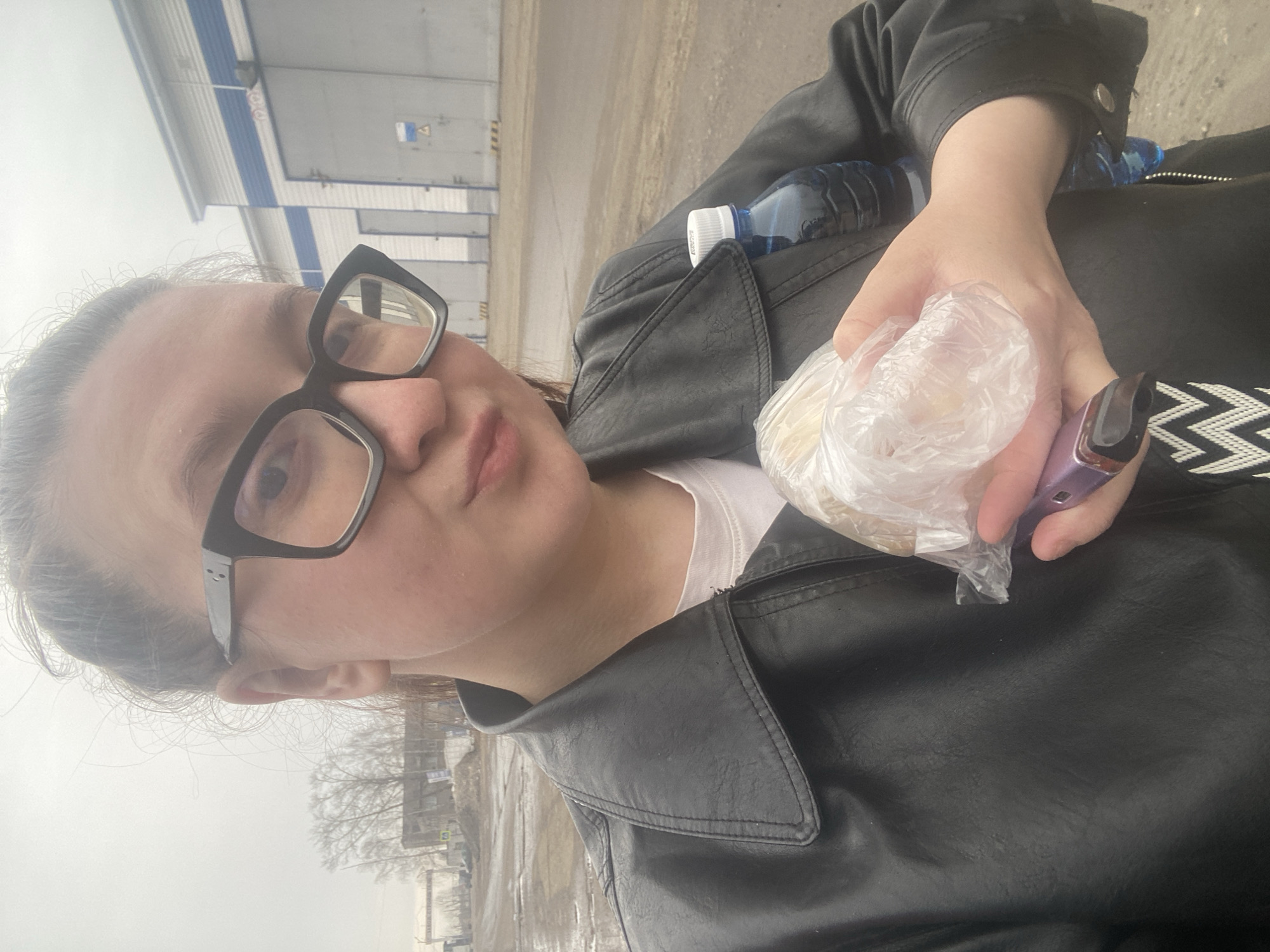 A woman wearing large black glasses and a black leather jacket is holding a small plastic bag containing food and a vaping device. A bottle of water is visible in her jacket. The background depicts an urban scene, likely in the city of Chaikovsky, Russia, characterized by industrial-looking buildings with loading docks and an overcast sky. The ground appears to be wet or slushy. A street with bare trees lines the background.