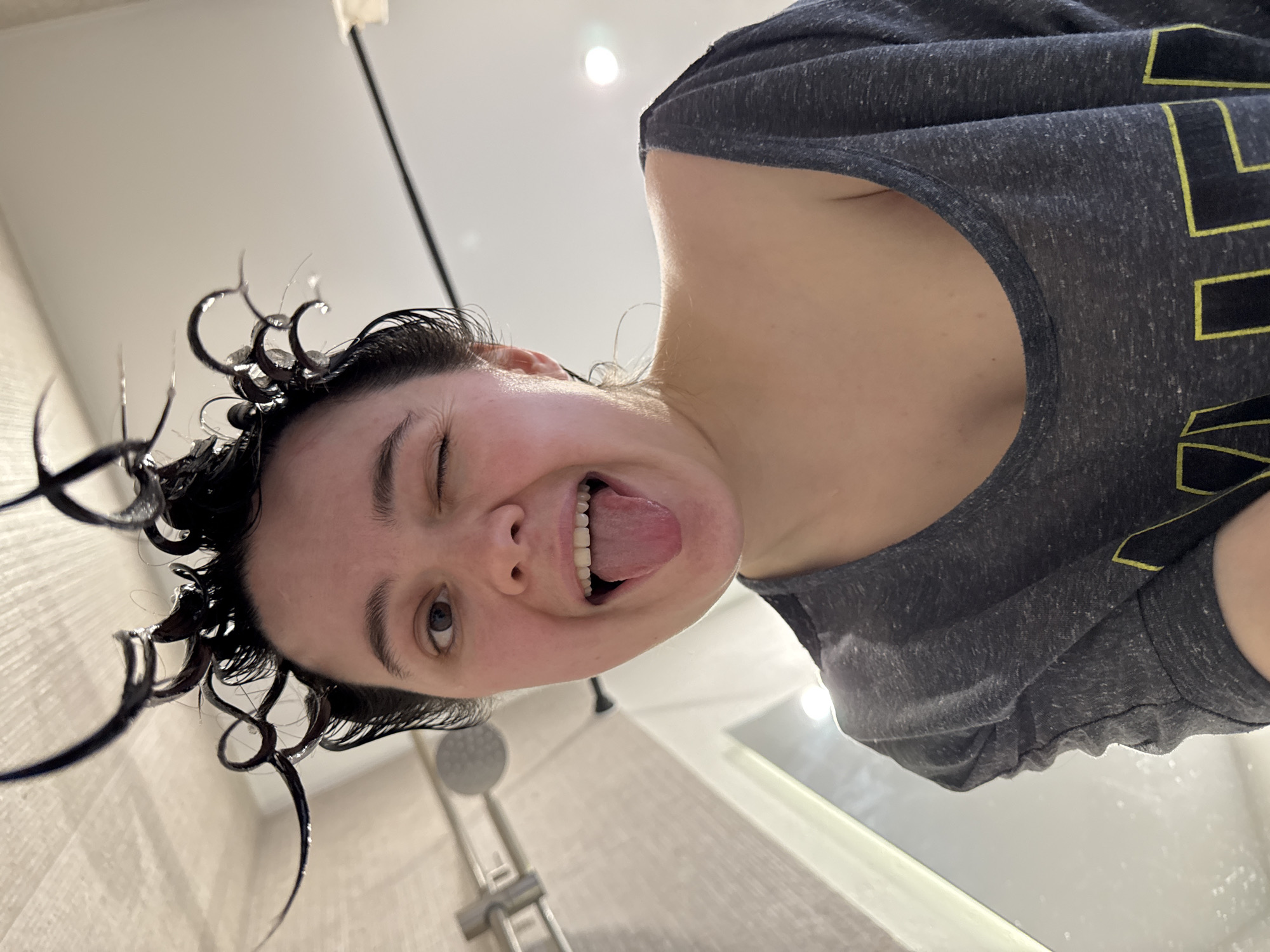 A low-angle, close-up photograph depicts a single female individual in a bathroom setting within Izhevsk, Russia. The subject, appearing to be in their 20s-30s, is cropped from the chest upwards and centered in the frame. Their dark hair is visibly wet, with several strands curled upwards and outwards, exhibiting water droplets. The individual's face is directed towards the camera, with their right eye (viewer's left) winking and their left eye open. Their mouth is open wide, revealing upper teeth, with the tongue fully extended. They are wearing a loose-fitting, dark gray, heathered short-sleeved top with a wide neckline, exposing the left shoulder. In the background, a silver-colored showerhead is partially visible on the left side of the frame. The ceiling is white, featuring a circular recessed light fixture in the upper right. A dark horizontal bar, possibly a shower frame element, spans the upper right background.