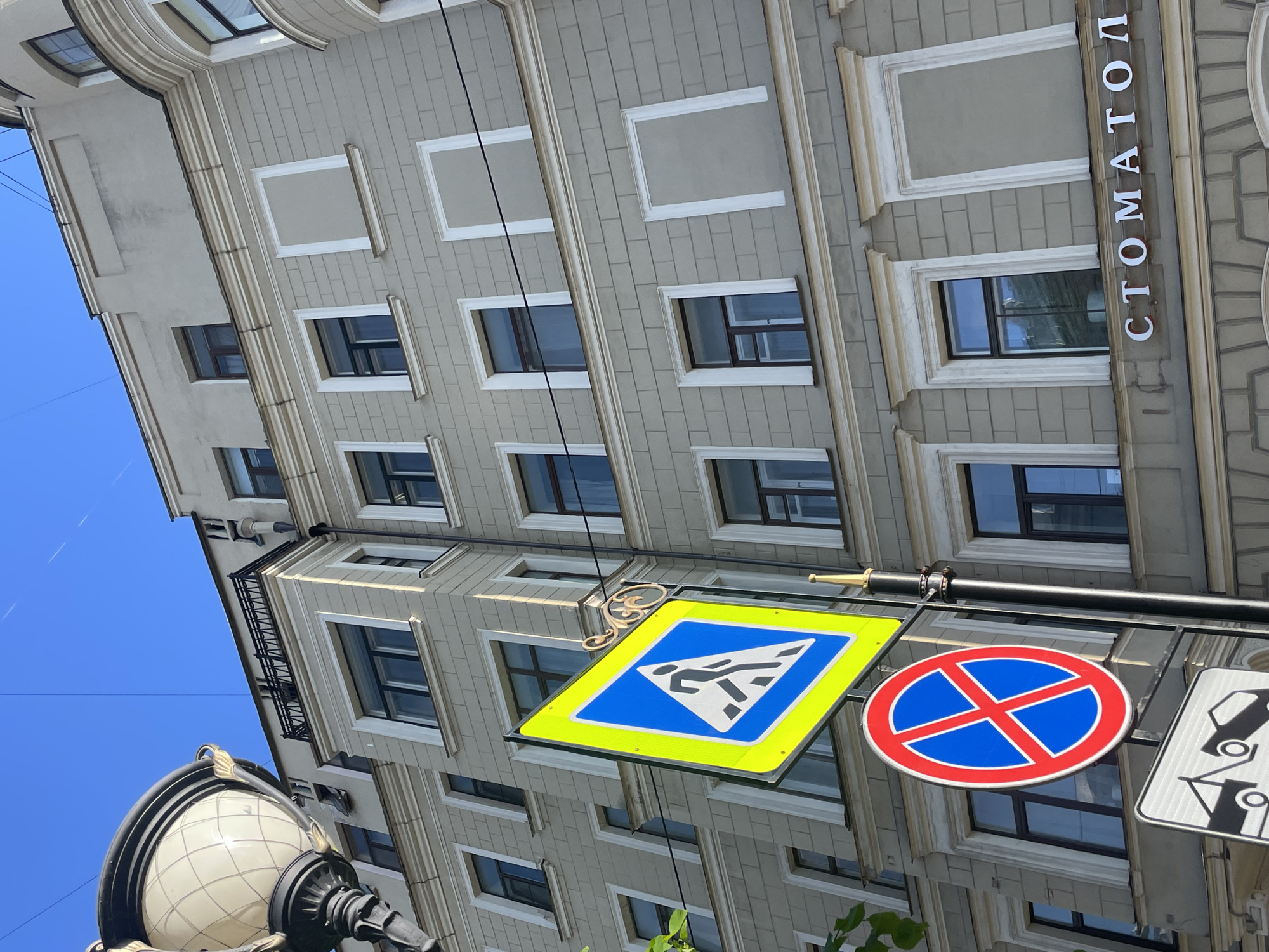 The image shows a street in Saint Petersburg, Russia, with a view looking up at a building and a streetlight with traffic signs. The building is light grey, with a rectangular facade. It has multiple floors, each with windows and decorative elements. The building has the word "СТОМАТОЛОГ" vertically printed on one side. In the foreground, there is a streetlight with a spherical lampshade and ornate metalwork. A horizontal bar is connected to the streetlight, holding traffic signs. These signs include a yellow and blue pedestrian crossing sign, a round red and blue "no parking" sign, and a third, partially visible sign. The sky is blue and clear.