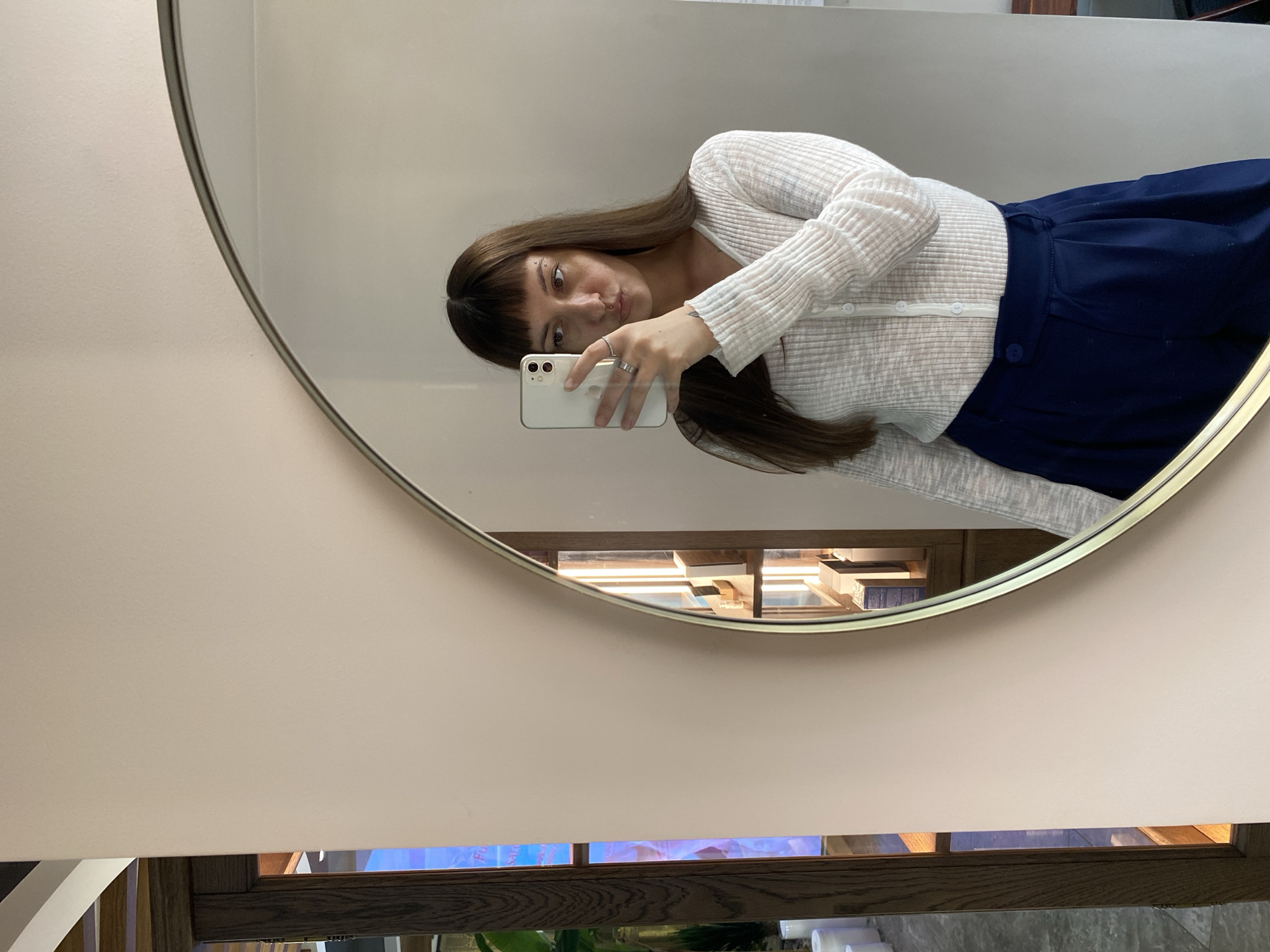 A woman takes a mirror selfie indoors. She is wearing a white, ribbed, long-sleeved shirt with buttons down the front and dark blue pants. She has bangs and long brown hair. Her left hand is holding a white smartphone up to take the picture, and she is wearing rings on her fingers. Her face is reflected in a large, oval mirror with a thin metal frame. To the left of the mirror, a wooden display cabinet with shelves and illuminated items is visible. The wall behind her is a pale pink color. The environment does not provide any specific clues to its location in Votkinsk, Russia.