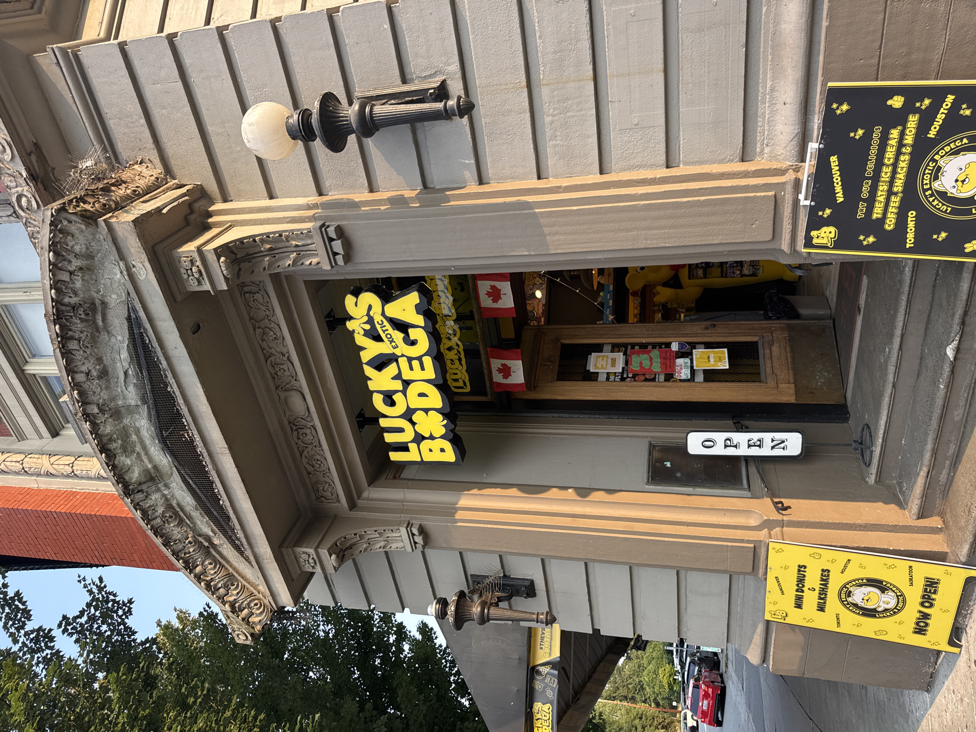 The image depicts the entrance to "Lucky's Exotic Bodega" in North Vancouver, Canada. The building features ornate architectural detailing above the doorway, including a stone archway with carved ornamentation. A neon yellow sign with black lettering reads "LUCKY'S EXOTIC BODEGA" and is illuminated. Two Canadian flags are hung inside the entrance. To the left of the entrance, there is a yellow A-frame sign advertising "MINI DONUTS & MILKSHAKES" and "NOW OPEN!" The sign also lists locations such as Vancouver, Toronto, Houston, and Saskatoon. To the right of the entrance, another A-frame sign, this one black with yellow lettering, advertises "TRY OUR DELICIOUS TREATS! ICE CREAM, COFFEE, SNACKS & MORE." A white "OPEN" sign on a stand is positioned on the bottom step. A vintage-style black lamppost with a white globe light is mounted on the building's exterior to the right of the entrance. The building's facade is clad in horizontal grey panels. A large green tree is visible to the left of the building, and a street with parked cars and an overhead structure (possibly a bridge or overpass) is in the background. The lighting suggests it is late afternoon or early evening, with sunlight casting long shadows.