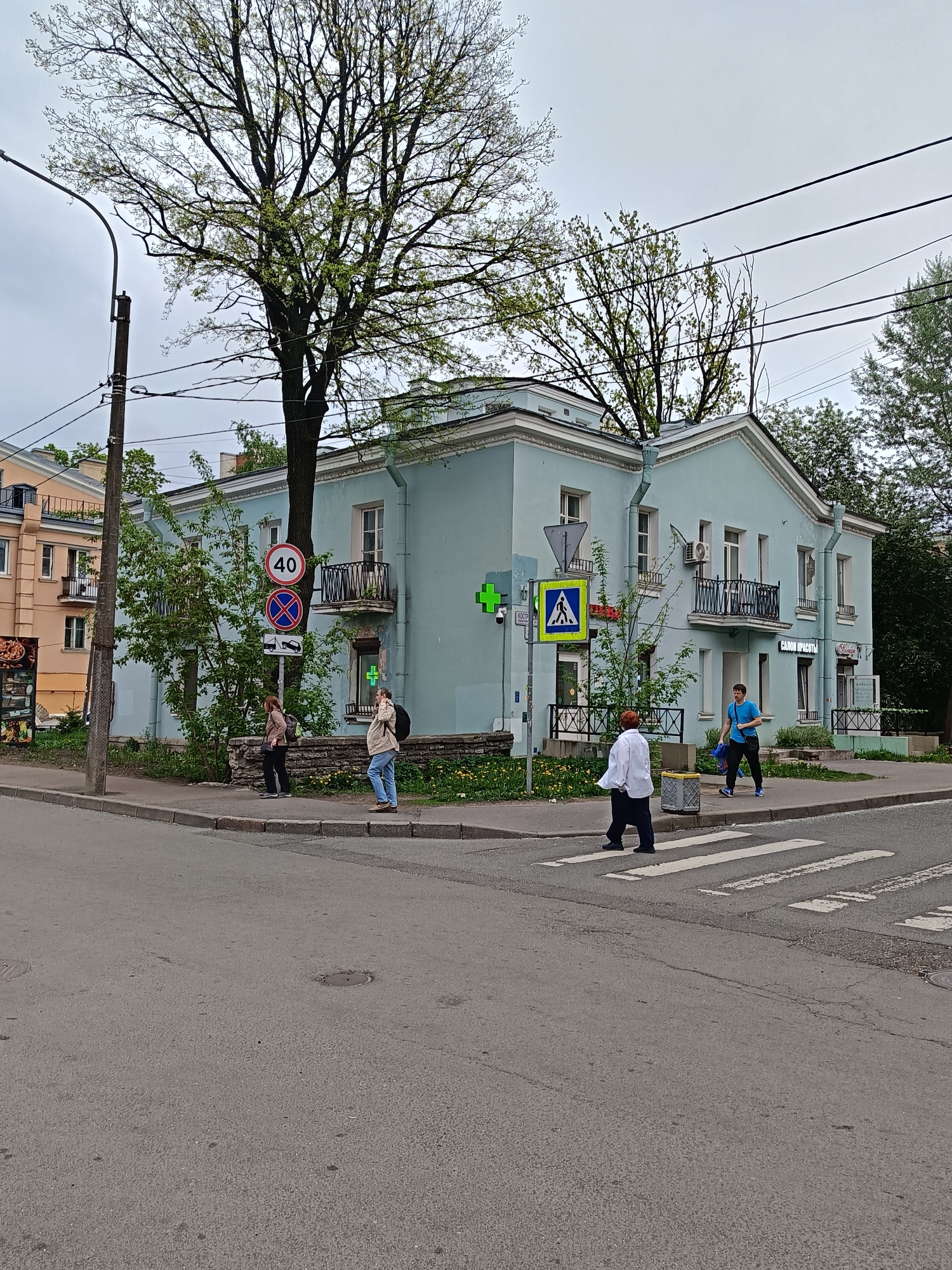The image shows a street scene in Saint Petersburg, Russia. An intersection with a crosswalk is visible, and several people are present. Three individuals are standing on the sidewalk near a road sign indicating a speed limit of 40 km/h. Another person is walking across the crosswalk, heading towards a man walking on the same crosswalk. The buildings in the background are a mix of colors and architectural styles, including a two-story light blue building with balconies and a pharmacy sign. Trees are scattered throughout the scene. The sky is overcast.