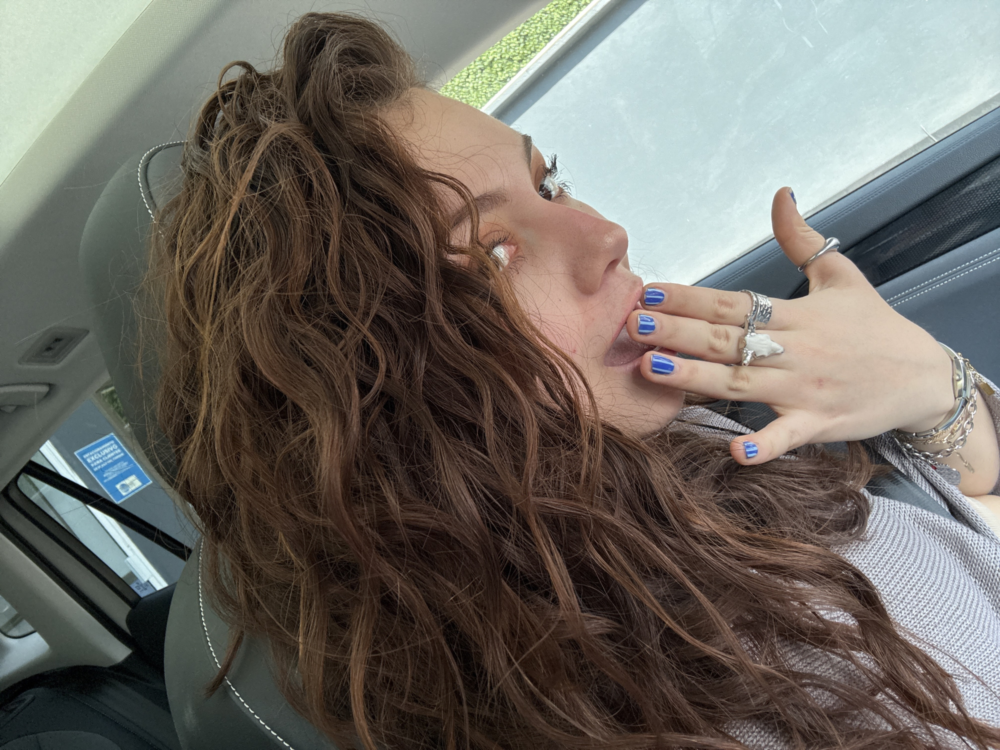 A side-view, medium shot depicts a fair-skinned young woman with curly, auburn hair piled loosely on top of her head. Her gaze is directed towards the upper right of the frame, and her open mouth is partially obscured by her left hand. Her fingernails are painted a vibrant blue. She is wearing multiple silver rings and bracelets. She appears to be seated in a car, as evidenced by the car seat and window in the background. A sticker is partially visible on the car window. The location appears to be Mexico City, Mexico, based on the language of the sticker.