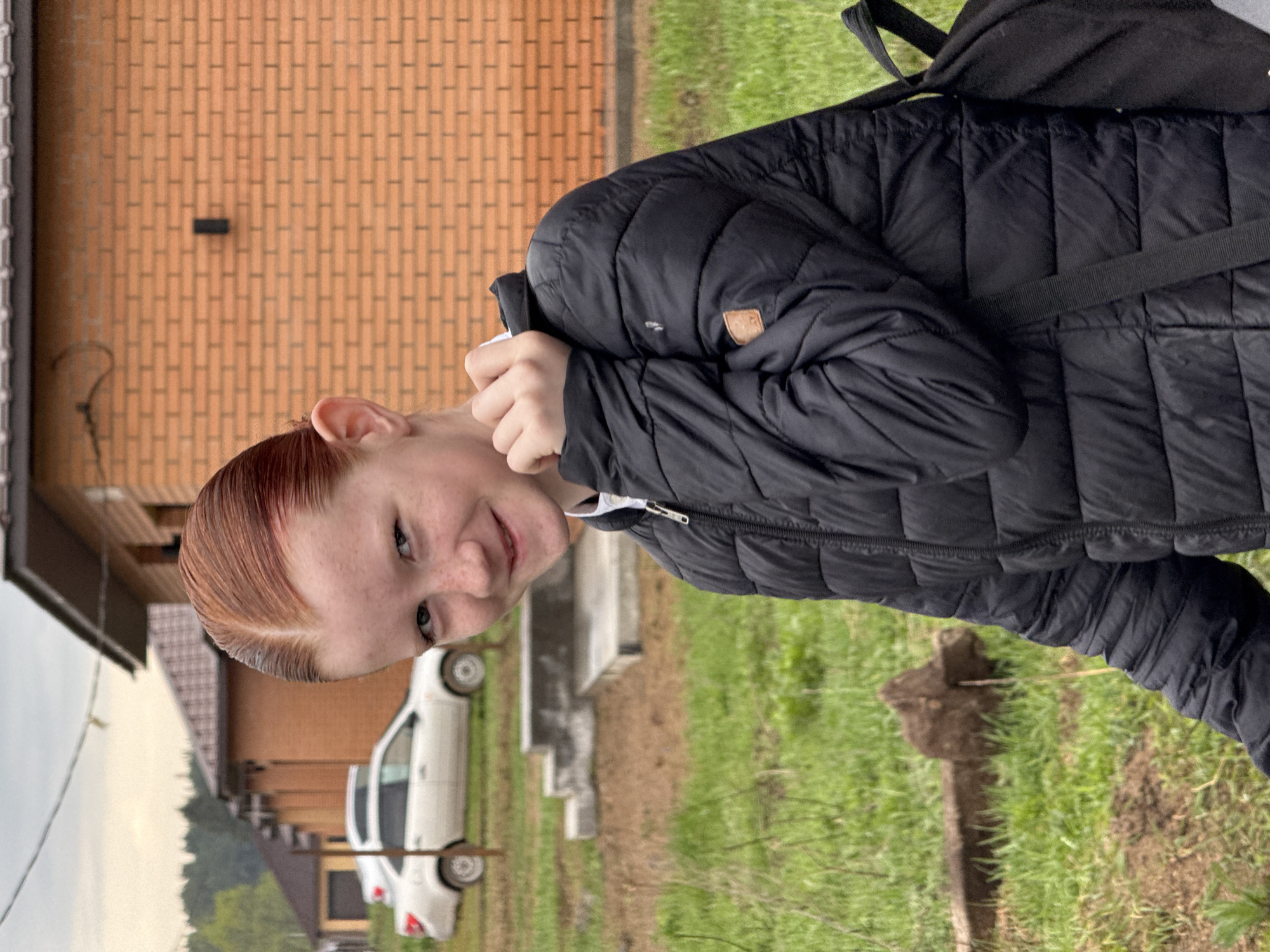 A person with reddish-brown hair, wearing a black puffy jacket with a backpack over one shoulder, is standing outside. In the background is a brick building and a white car parked near a house with a visible roof. The person is standing on grass. The location is not discernible as Chaikovsky, Russia from visual cues.