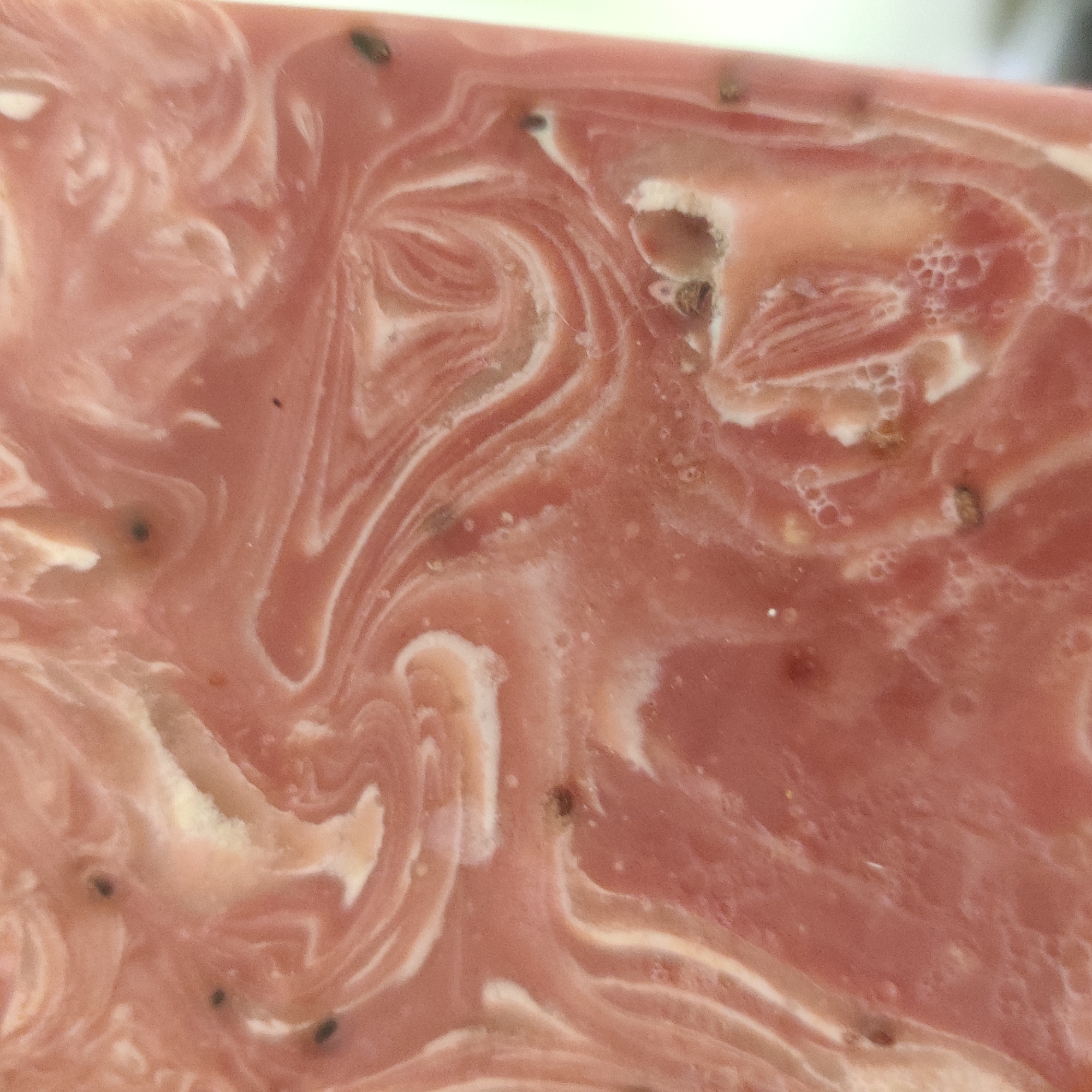 The image is a close-up, high-angle shot of a pink and white swirled substance, likely soap or a similar material. There are small, dark specks embedded within the substance, appearing to be seeds or exfoliants. The texture is smooth with some areas showing small bubbles or imperfections. The lighting suggests an indoor environment, but no specific location or context within Yekaterinburg, Russia, can be determined from the visual information provided.
