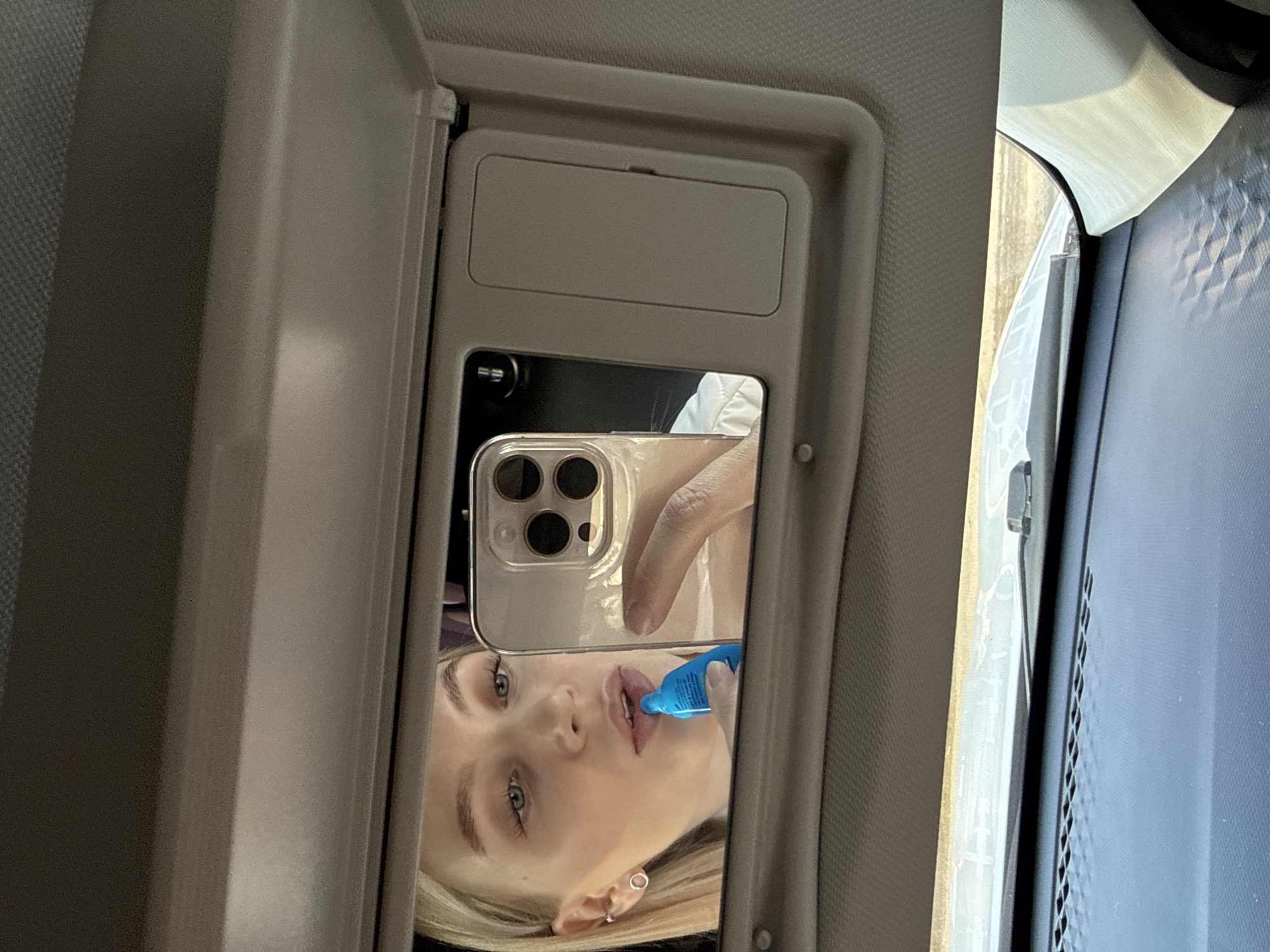 A selfie is being taken in a car vanity mirror. The subject is a blonde woman with fair skin who is applying a blue substance, possibly lip gloss, to her lips. The phone being used to take the photo has a clear case and multiple camera lenses. The car's interior, including the vanity mirror and adjacent surfaces, is visible. The location is not evident based on this image, and therefore cannot be identified as Izhevsk, Russia.