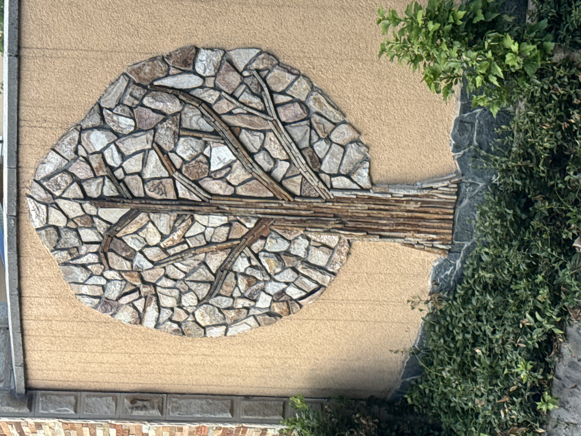 The image shows an outdoor wall decorated with a mosaic of a tree. The tree mosaic is composed of various natural stones, with a trunk made of vertical, slender stones and a crown formed by irregularly shaped stones in shades of grey, beige, and brown. The wall itself is textured, painted a light beige color, and appears to be part of an exterior building facade or a decorative garden wall. To the left and right of the tree mosaic, there is green foliage, suggesting a garden or landscaped area. A stone border runs along the base of the wall, and a segment of a brick wall is visible on the far left. The overall impression is of a decorative art installation on a wall. The location is identified as being in the city of Ialoveni, Moldova.