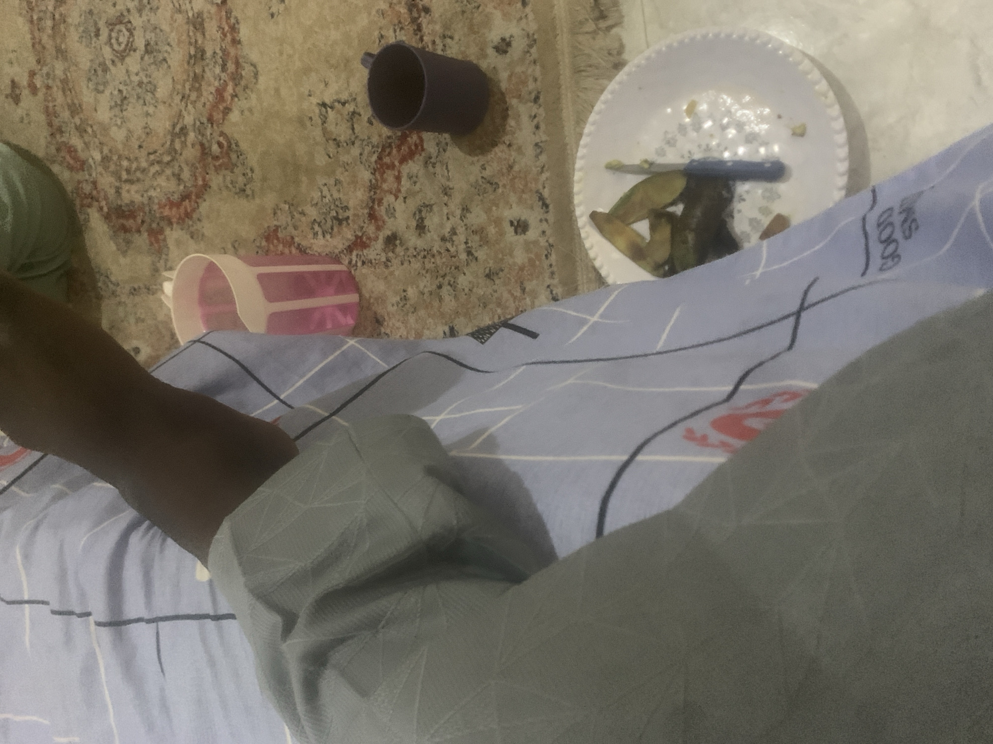 The image depicts a close-up view of a person's arm and hand resting on a light blue and white striped fabric, possibly a bedsheet or blanket. The fabric has a subtle patterned texture and some visible red markings that appear to be part of a logo or design. In the background, a patterned carpet is visible, with a pink and white cup filled with a pink liquid to the left of the person's arm. To the right of the arm, a dark purple mug sits on the carpet. Further to the right and slightly lower, a white plate contains pieces of what appears to be cooked food, possibly eggplant, along with a blue-handled knife. The overall scene suggests a casual setting, perhaps indoors in a residential area. The location is specified as Kuson, Nigeria.