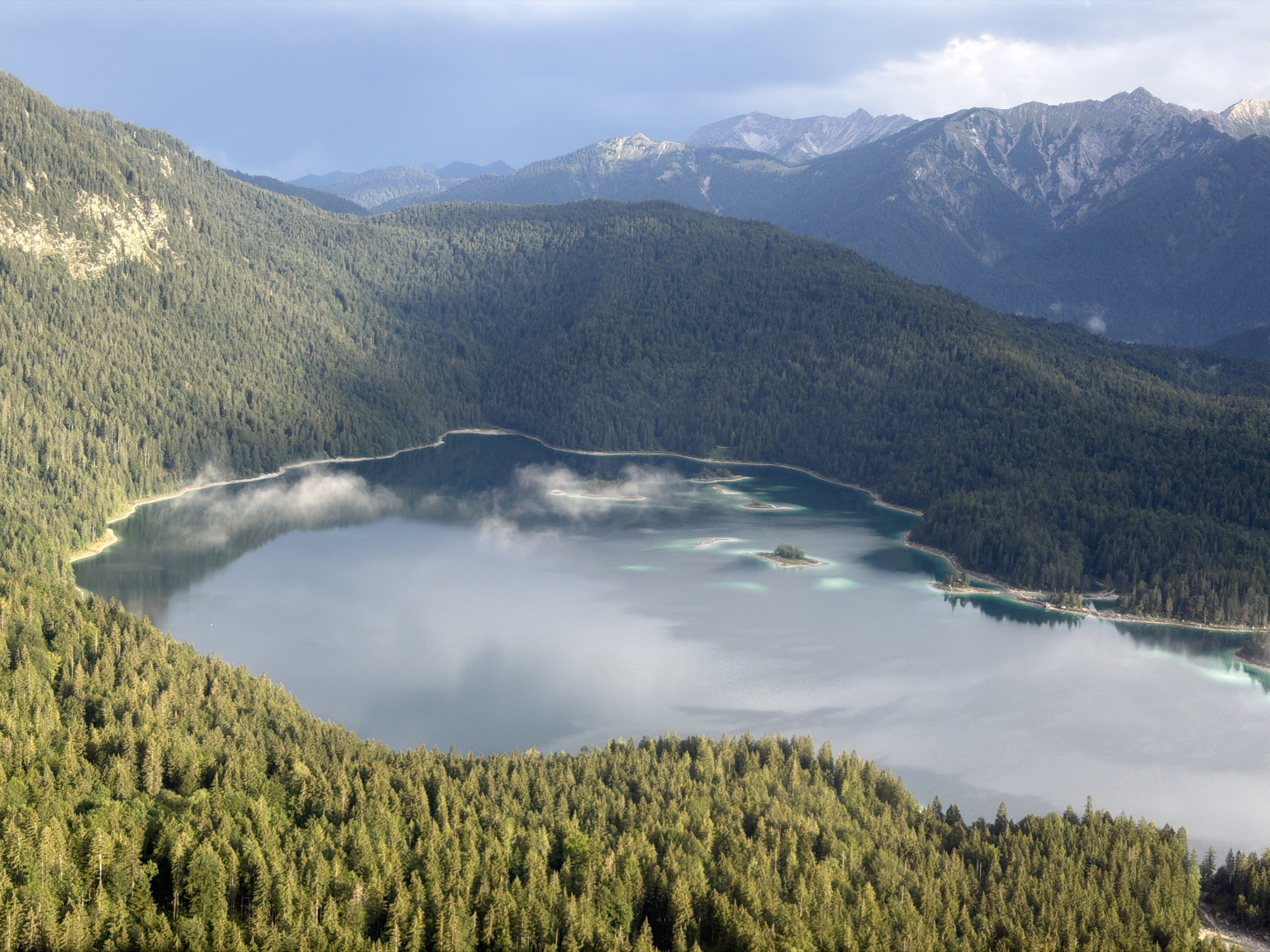 This is an aerial, high-angle panoramic view of the Eibsee lake near Grainau, Germany. The lake is a prominent turquoise color with several small islands scattered throughout. Mist hangs low over parts of the water. The lake is surrounded by dense, dark green coniferous forests that climb up the steep slopes of surrounding mountains. The mountains in the background are rocky and rugged, with some peaks showing patches of snow. The sky above is a mix of blue and grey clouds. The scene is natural and serene, with no signs of human activity.