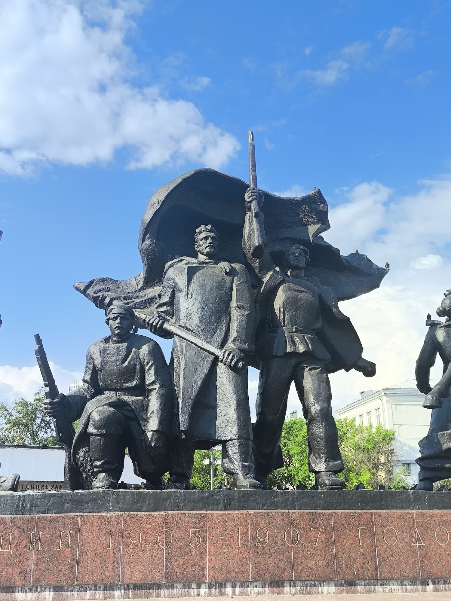 The image shows a bronze statue of a group of people on a stone base, set against a blue sky with scattered clouds. The statue depicts three prominent figures in the center, with what appears to be supporting figures on either end. The inscription on the base reads "1905-1907". Two birds appear to be resting on top of the flag.
Based on these details, this is likely a monument to the 1905-1907 Revolution, located in Moscow, Russia.