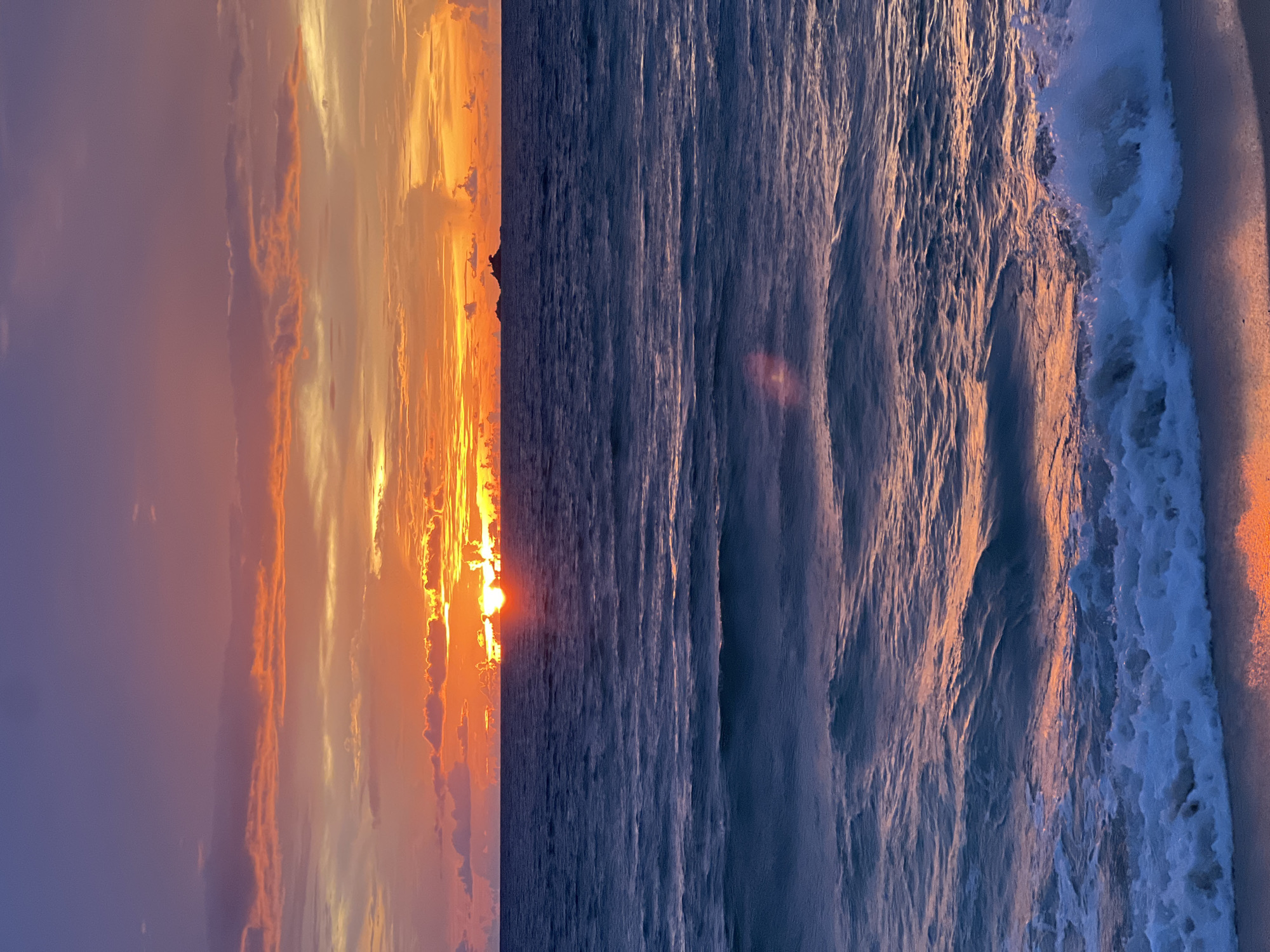 This media file depicts a serene sunset over the ocean. The scene is split vertically, with the left side showcasing a dramatic sky filled with vibrant orange and yellow clouds, illuminated by the setting sun. The right side captures the vast expanse of the deep blue ocean, with gentle waves rolling towards the shore. The sunlight, reflecting off the water's surface, creates shimmering patterns and casts a warm glow on the ocean. A small patch of land is visible in the distance, silhouetted against the bright sky. The atmosphere is peaceful and tranquil, evoking a sense of calm and awe. The time of day is clearly sunset, given the colors of the sky and the low angle of the sun. The weather appears clear and pleasant, with the ocean waves suggesting a calm sea. The location is a coastal area, likely a beach, though specific geographical cues are absent. There is no visible text in the media file.