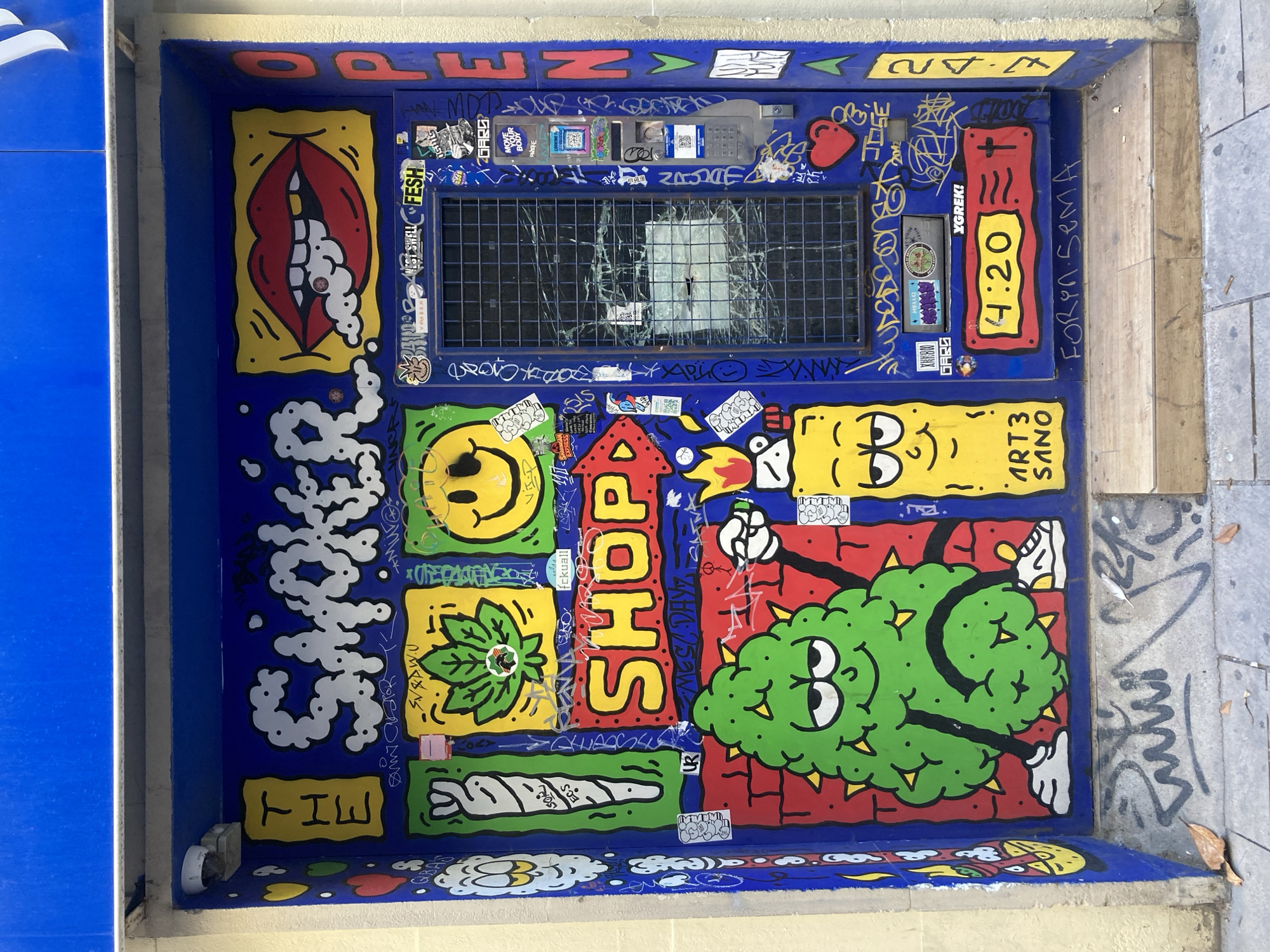 The image shows a blue wall with a variety of cartoonish graffiti art in what appears to be a small alcove. The art includes imagery related to cannabis, such as a large green bud character, a rolling paper, and a marijuana leaf. There are also images of a mouth exhaling smoke, a smiley face, and the word "SHOP" in red and yellow. A rectangular window with a metal grid is visible towards the top, and it is broken. The surrounding wall is also covered in stickers and tags. To the left is a blue wall and to the right is the sidewalk. The text "THE SMOKER" is displayed prominently in white letters. The text "4:20" is visible, and the graffiti art includes a reference to "ART3 SANO." Based on the presence of the text "FORON SemA" and the overall aesthetic, it's likely that this is located in Barcelona, Spain.