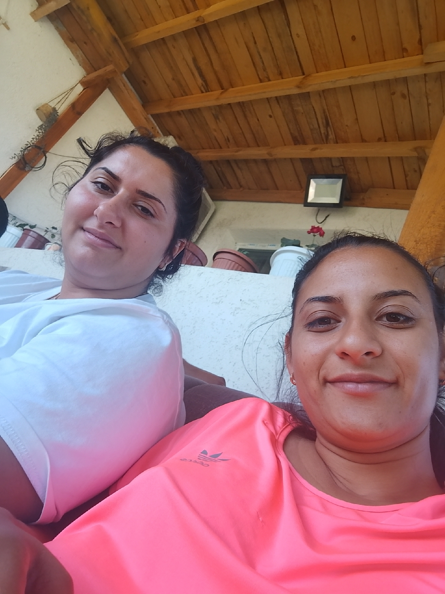 In Zimnitsa, Bulgaria, two women are captured in a selfie. The woman in the foreground, wearing a bright pink t-shirt with an Adidas logo, smiles warmly. Behind her, another woman in a white t-shirt gazes at the camera. They are seated outdoors under a wooden-beamed roof. In the background, a light fixture is mounted on the ceiling, and several potted plants are visible. The scene suggests a casual, relaxed moment.
