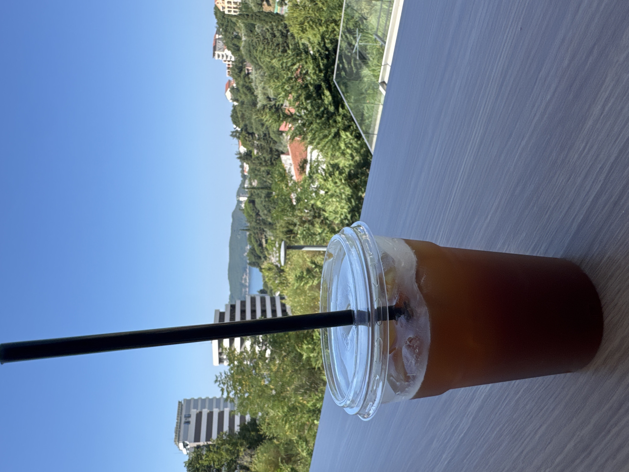 This is a vertical, eye-level shot of a clear plastic cup filled with a dark, iced beverage, likely coffee or tea. The cup has a clear plastic lid with a straw inserted. The beverage is amber to dark brown in color and contains ice cubes. The cup sits on a light-colored, textured surface that appears to be a table or deck railing.
In the background, a lush green hillside is dotted with buildings, suggesting an urban or resort area built into the landscape. The buildings are a mix of white and light-colored structures with some red-tiled roofs visible among the trees. The sky is a clear, bright blue, indicating a sunny day. The overall scene suggests a leisurely outdoor setting, possibly a cafe or balcony with a view. The time of day appears to be daytime, with bright sunlight casting shadows.
The specific location context is Boreti, Montenegro. The presence of multiple buildings on the hillside and the view towards what could be the sea in the distance are consistent with a coastal Montenegrin setting. There is no visible text in the image. The overall mood conveyed is one of relaxation and enjoyment of a beverage in a scenic environment.