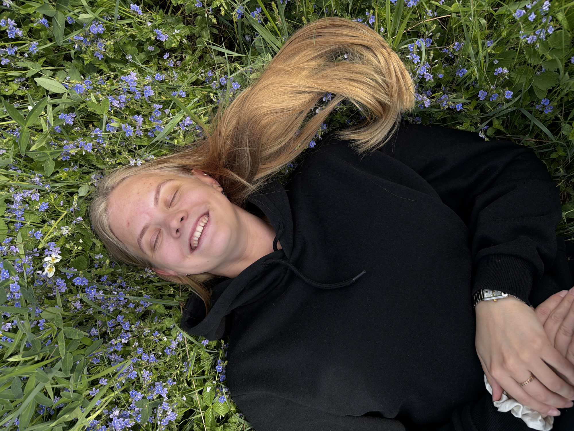 A young woman with blonde hair is lying on a field of green grass interspersed with small blue flowers, likely in a park or meadow. She is wearing a black hoodie and a silver wristwatch. She is smiling with her eyes closed, appearing relaxed. Her hands are clasped together. The exact location within Chaikovsky, Russia is not discernible without further information.