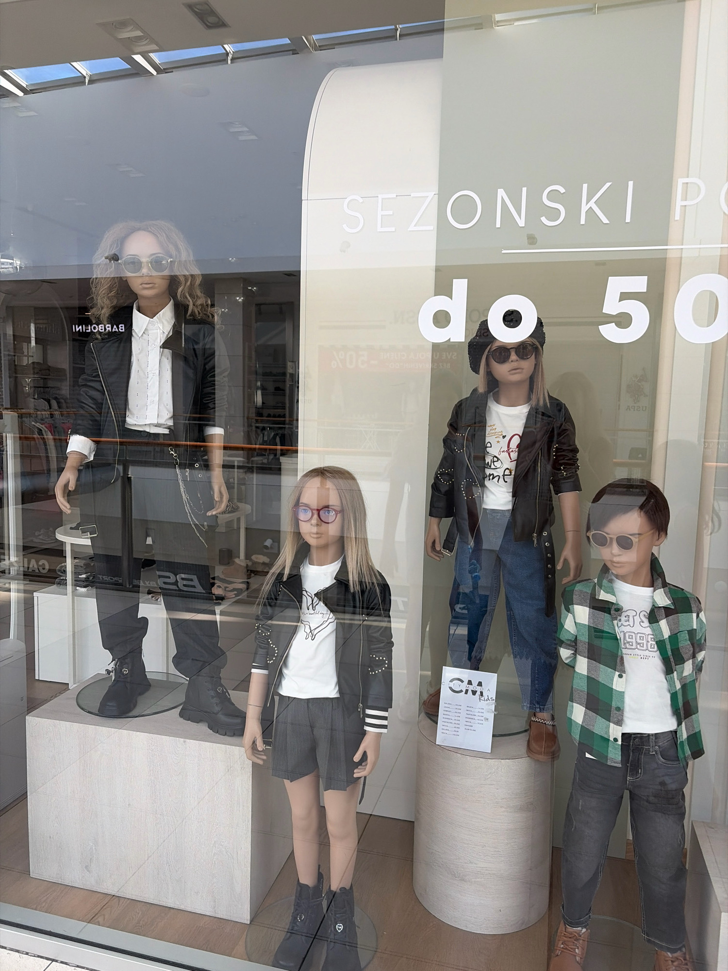 This image shows a storefront window display featuring four mannequins dressed in casual clothing. The mannequins are arranged in a staggered formation, with two adult-sized mannequins in the back and two child-sized mannequins in the front. The display is illuminated by overhead lighting and reflects the surrounding environment, suggesting it is daytime.
On the left, a taller adult mannequin wears a white collared shirt, a black leather jacket, and dark pants. It has curly blonde hair and is wearing sunglasses. To its right, a child-sized mannequin is dressed in a black leather jacket with studded details, a white t-shirt with a graphic print, blue jeans, and brown boots. This mannequin is also wearing sunglasses and a black beret. In the center right, another child-sized mannequin wears a green and black plaid shirt over a white t-shirt, dark jeans, and brown boots, also sporting sunglasses. The mannequins are positioned on display stands and pedestals.
A sign is visible on a cylindrical pedestal in front of the child mannequin on the right, displaying "CM kids" and a list of text, likely prices or product information. In the background, through the glass, clothing racks and shelves are faintly visible, indicating the display is part of a retail store. Text on the glass in large white letters reads "SEZONSKI PO" and "do 50", suggesting a seasonal sale with up to 50% off. The overall impression is a modern fashion store catering to both adults and children. The reflection in the glass suggests the store is located in an indoor shopping mall or a similar commercial setting, possibly in Podgorica, Montenegro, based on the context provided for this query.