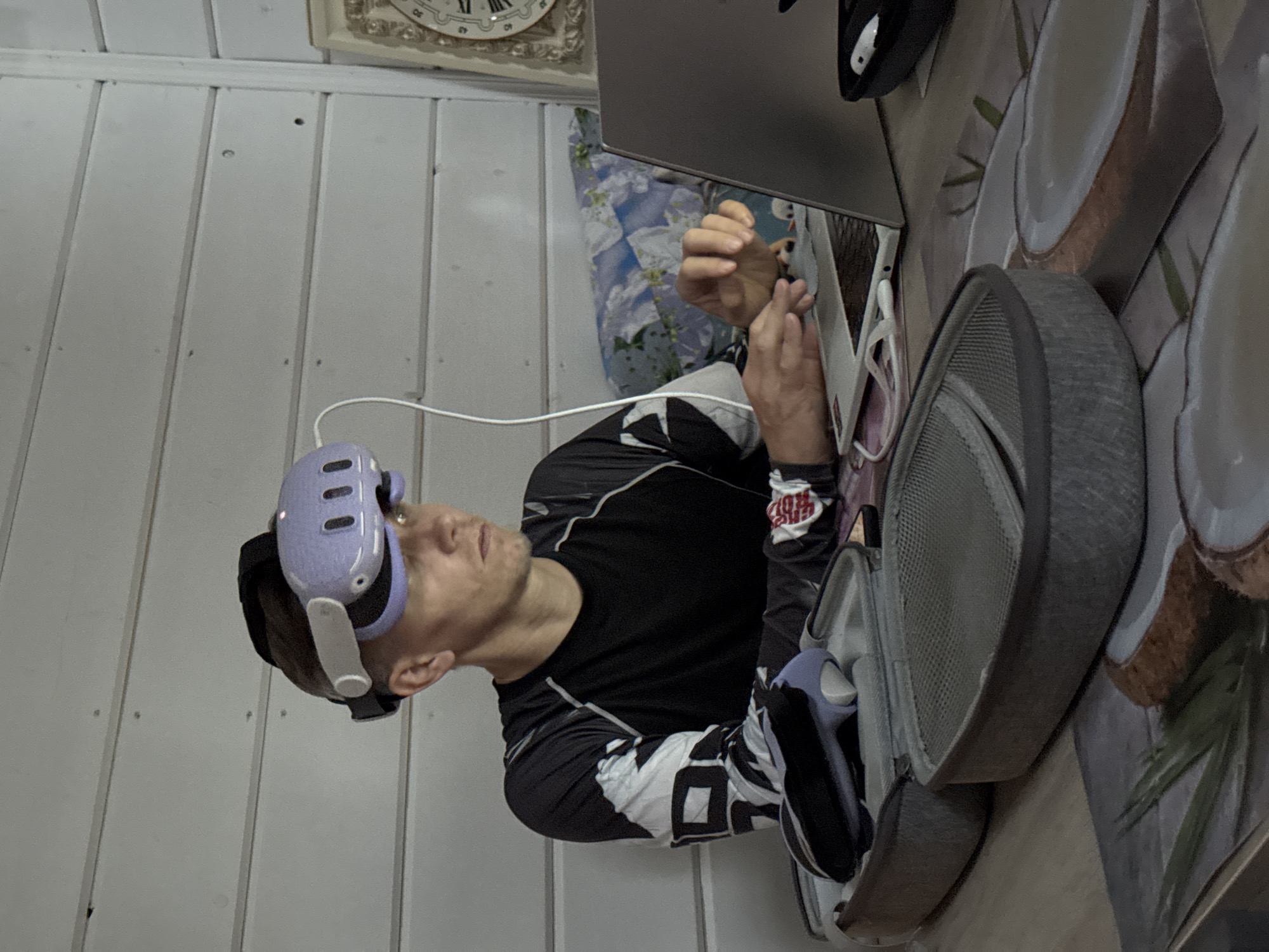 In the city of Zelenaya Roshcha, Russia, a man with short hair is seated at a table, wearing a light purple virtual reality headset. He is looking towards a laptop situated on the table in front of him, with his hands positioned near the keyboard. The man is wearing a long-sleeved black and grey shirt with a graphic design on the sleeves. Beside the laptop is a grey protective case, open, revealing a controller and other accessories. The table surface has a printed pattern featuring coconuts and palm leaves. In the background, a white wooden paneled wall is visible, with a framed clock on the right side. A floral patterned pillow rests against the wall behind the man. The environment appears to be indoors, possibly a home or a casual workspace.