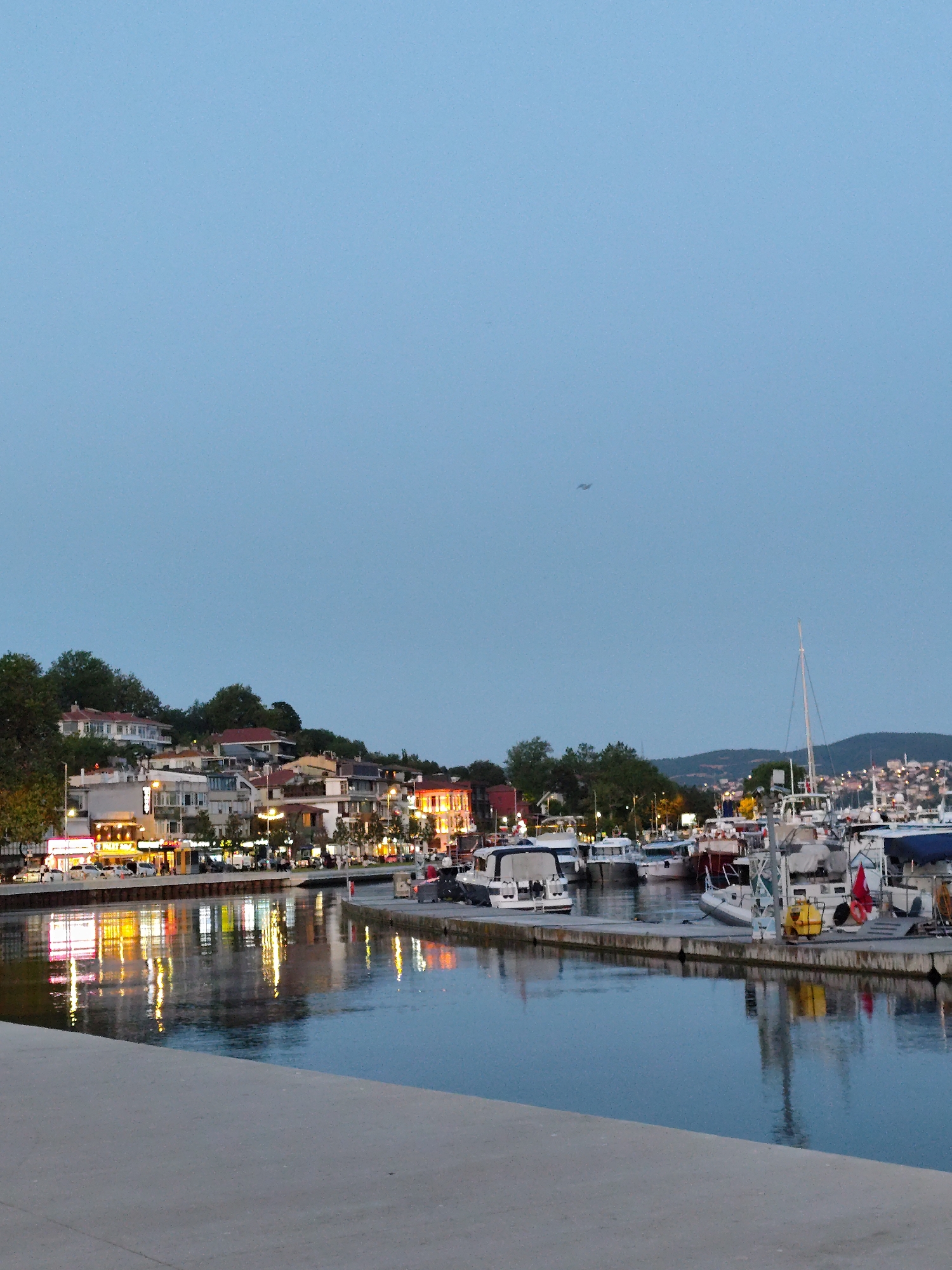 In the city of Sarıyer, Turkey, a waterfront scene unfolds at dusk or early evening. The foreground is dominated by a concrete embankment, possibly a walkway or pier, sloping down towards the water. The calm, dark water of a harbor or bay reflects the illuminated buildings and lights from the opposite shore. These buildings are situated on a gentle incline, with trees and foliage interspersed among them. The architecture appears to be a mix of residential and commercial structures, with some displaying bright, warm-toned lights, indicating shops or restaurants.
Across the water, a fleet of boats is docked. These range from small motorboats to larger vessels, some with white covers or canopies. A prominent sailboat with a tall mast stands out among the others. A red flag, possibly the Turkish flag, is visible on one of the boats. The sky above is a pale blue, transitioning towards twilight, with a hint of a distant hillside or mountain range visible on the far horizon, also dotted with lights. A small, dark speck, potentially a bird or aircraft, is visible in the upper portion of the sky. The overall impression is one of a serene, yet active, evening at a coastal town.