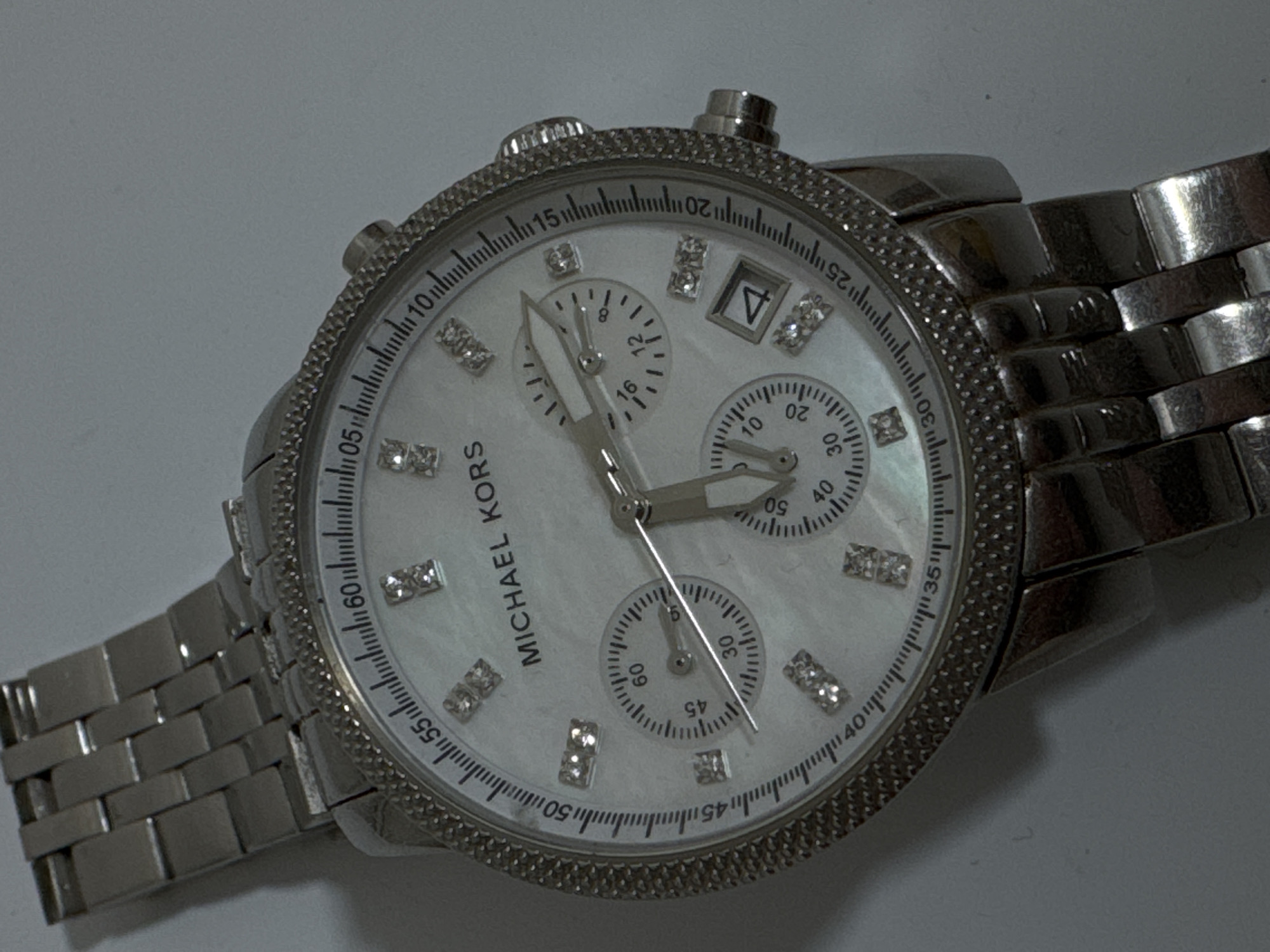 An overhead, slightly angled shot shows a silver Michael Kors watch with a white mother-of-pearl face, lying on a gray surface. The watch has a metal band and a bezel encrusted with small, clear rhinestones. The face of the watch features diamond-shaped hour markers and three subdials. The main hands indicate a time of approximately 10:14. The date window shows the number 4. The watch is the sole subject of the image.