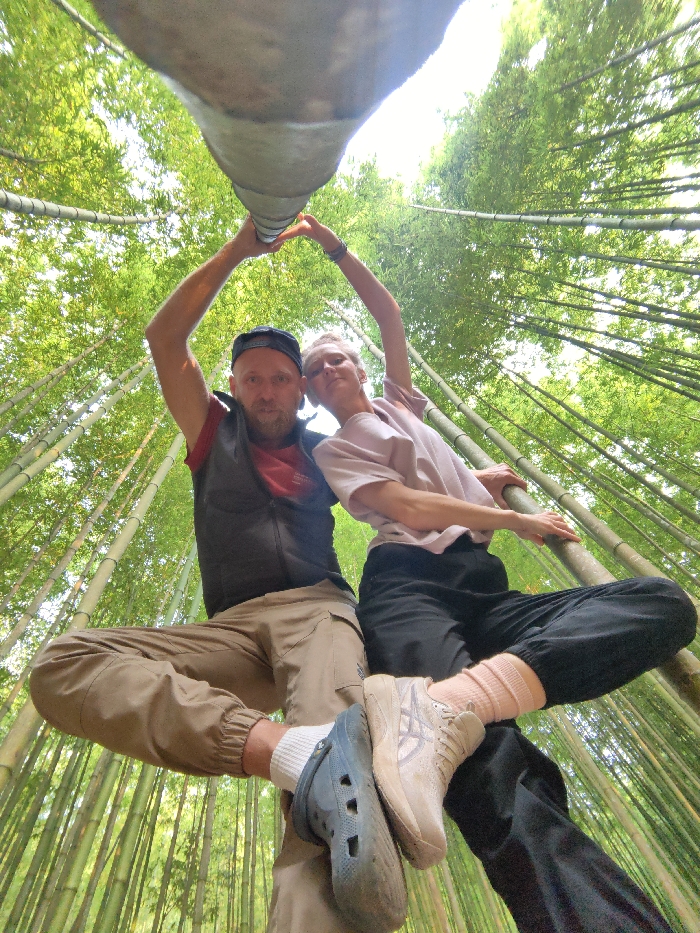 A wide-angle, low-angle photograph depicts a male and a female individual situated within a dense bamboo forest in Mù Cang Chải, Vietnam. The scene is dominated by numerous tall, green bamboo stalks ascending vertically into a bright sky. Centrally, the two individuals are positioned beneath a thick, pale cylindrical object, possibly a horizontal bamboo pole, that spans the upper portion of the frame.
The male, on the left, has a short beard and wears a dark baseball cap, a dark sleeveless vest over a red shirt, and light brown cargo pants. His left foot, wearing a blue Croc-style clog over a white sock, is visible. The female, on the right, has short, light-colored hair and wears a light pink short-sleeved top with loose-fitting black pants. Her right foot wears a light beige sneaker over a light pink ribbed sock. Both individuals have their arms raised above their heads, joining hands to form a heart shape beneath the central cylindrical object. The male's left foot is positioned beneath and slightly behind the female's right foot. The female's left arm extends horizontally to brace against a slender bamboo stalk to her right. Both individuals are looking directly towards the camera, which is positioned below them, facing upwards.