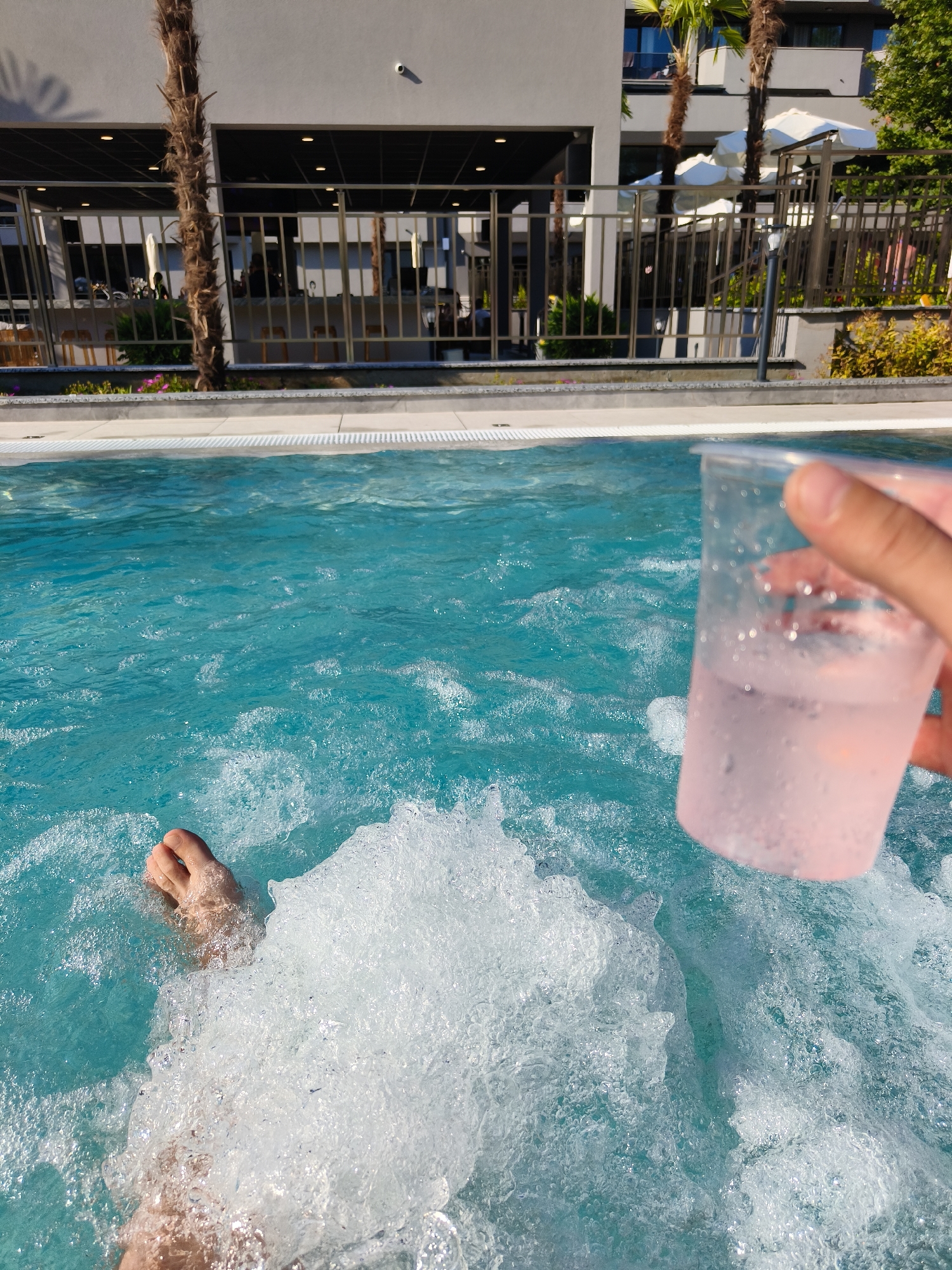 A first-person perspective view of a person relaxing in a bubbling outdoor swimming pool in Kiten, Bulgaria. The water is a vibrant turquoise and filled with frothy bubbles. In the foreground, a person's bare foot is visible partially submerged in the water on the left side of the frame. On the right side, a person's hand holds a clear plastic cup filled with a light pink liquid and ice, with condensation visible on the outside of the cup. In the background, a modern building with a dark awning, palm trees, and a metal fence are visible. A security camera is mounted on the building's exterior wall. The setting appears to be a resort or hotel with an outdoor bar or seating area beyond the fence. The sun is shining, casting shadows from the palm trees.