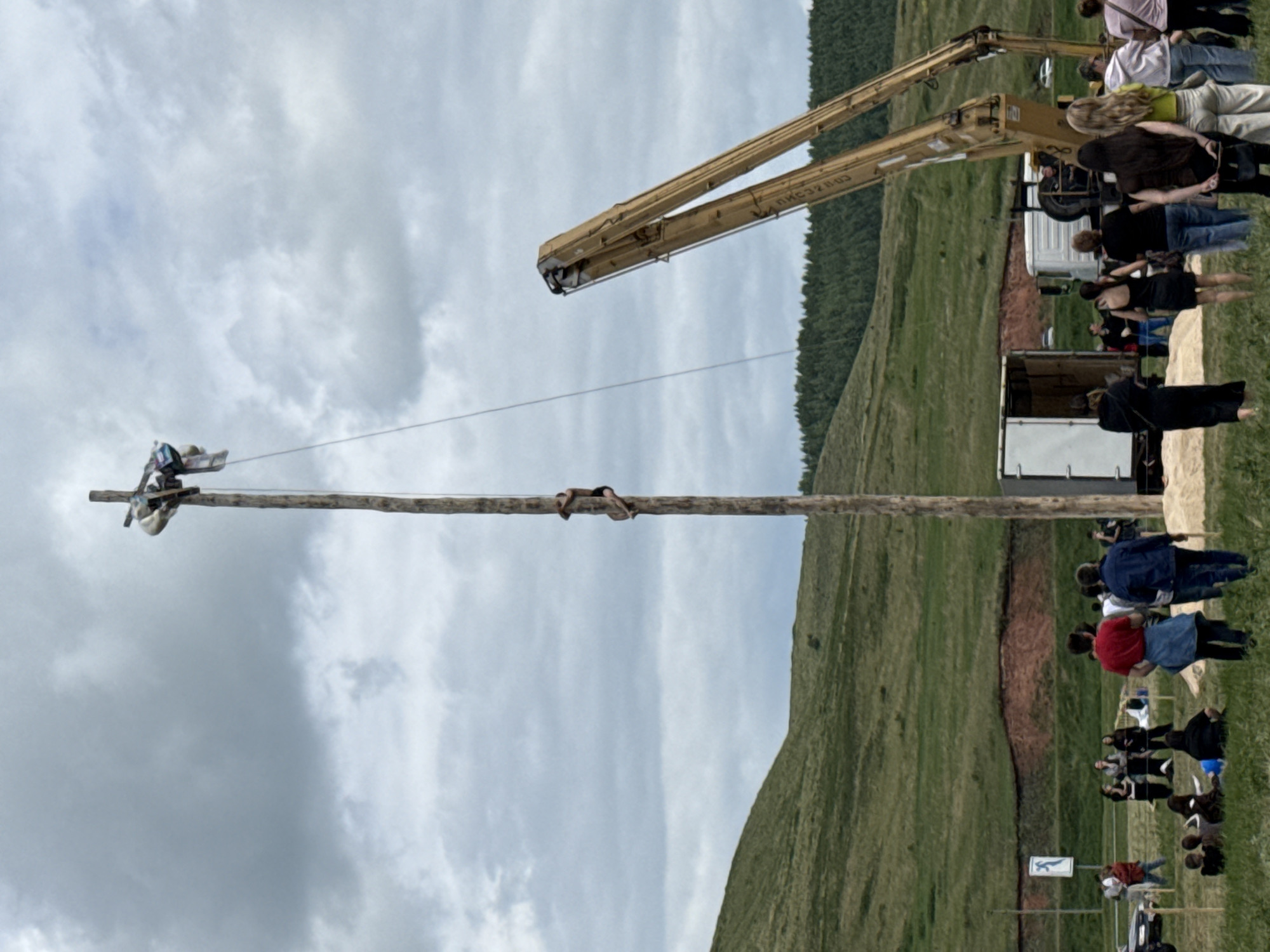 This image depicts an outdoor event in Nurykuevo, Russia, under a cloudy sky. The central focus is a tall wooden pole, at the top of which two individuals are positioned. One person is actively climbing the pole, clinging to it with their arms and legs. Another person is near the top, holding onto a cross-like structure. A rope extends from the top of the pole downwards.
In the foreground, a crowd of spectators watches the event. People are scattered across a grassy field, some seated, others standing. Several individuals are dressed casually, indicating a relaxed, community gathering.
Behind the crowd, a large yellow crane is visible, its arms extended. Nearby, a white truck and a smaller white vehicle are present, suggesting logistical support for the event. The background features rolling green hills and a dense line of trees. The overall scene suggests a local festival or competition involving pole climbing.