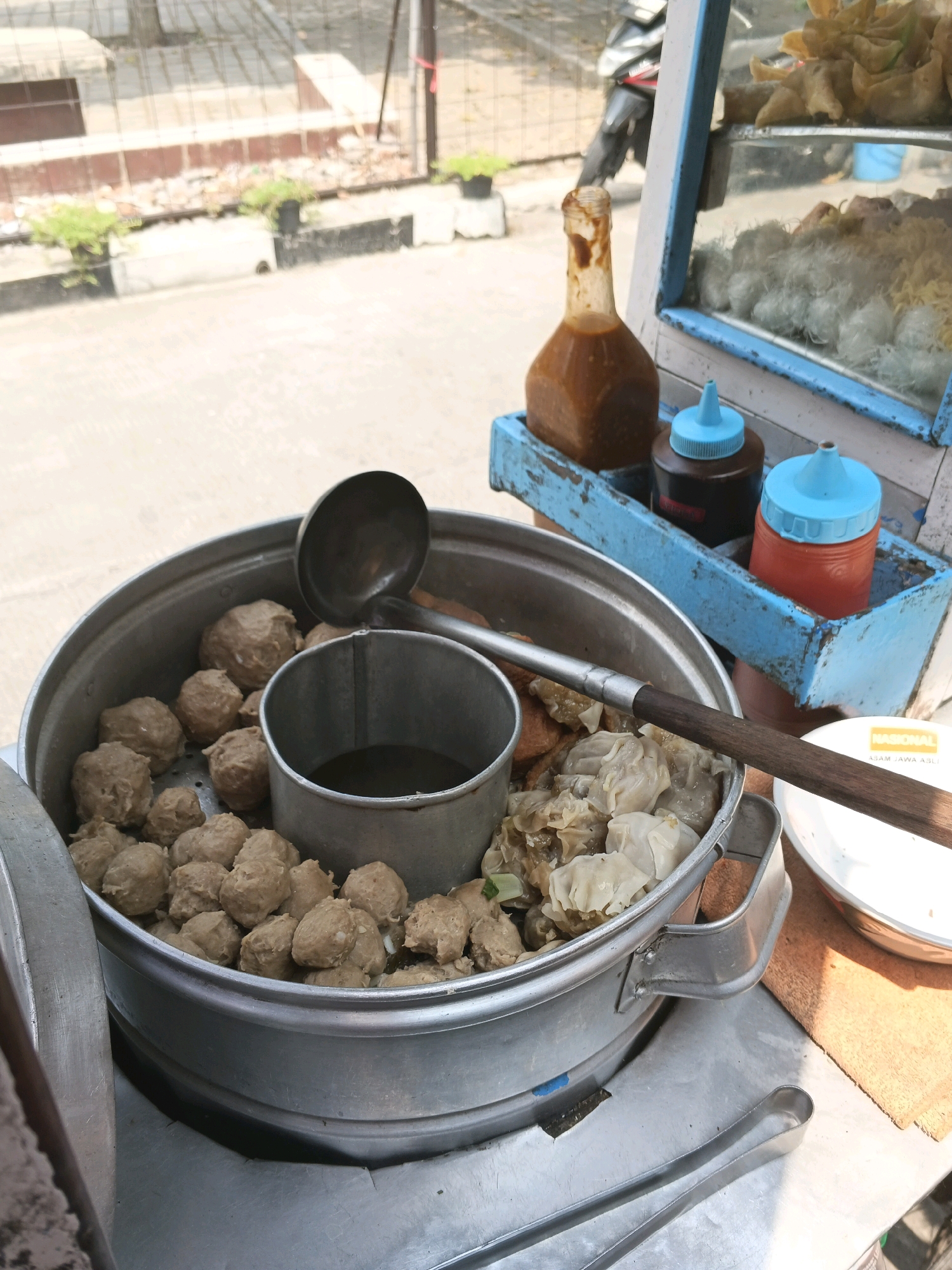 The image depicts a close-up view of a food cart, likely in Bandung City, Indonesia, selling Indonesian meatballs (bakso) and dumplings. A large, round metal pot filled with meatballs is prominently featured. A metal ladle rests on the edge of the pot, and a smaller metal container with broth is visible inside. Adjacent to the meatballs are steamed dumplings. The food cart has a blue-trimmed display case containing more food items. Two condiment bottles, one with red liquid and the other with dark brown liquid, are placed in a blue holder. The cart is positioned outdoors, with a street and some foliage visible in the background. The overall lighting suggests a sunny day. No people are visible in the frame.