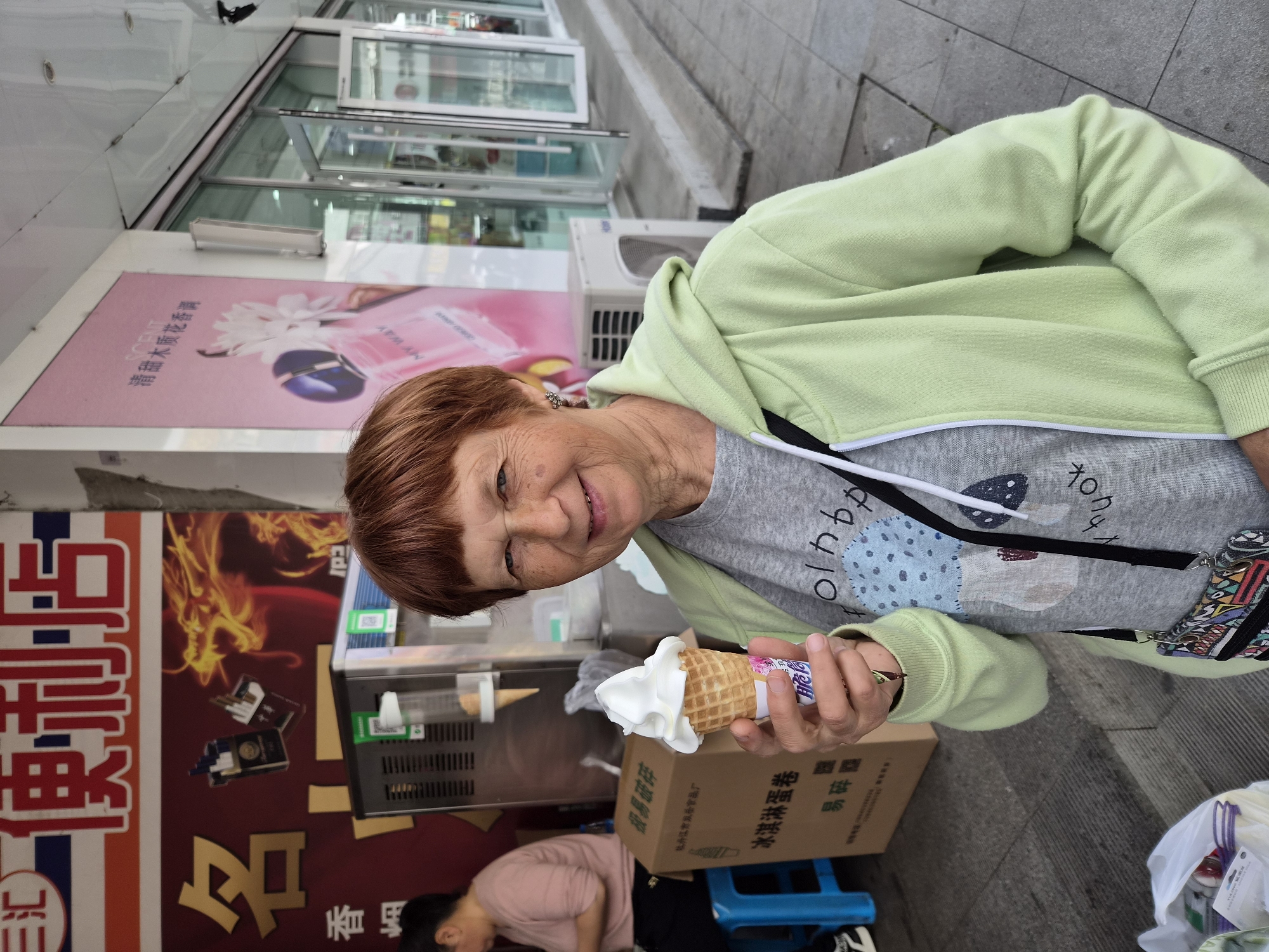 An elderly Caucasian woman with short red hair and glasses is holding a vanilla ice cream cone in her right hand, smiling at the camera. She is wearing a light green zip-up hoodie over a grey t-shirt that has a graphic print of mushrooms. A black strap, likely for a bag, is visible across her chest. In the background, a storefront with glass doors and windows is visible, along with signage in Chinese characters. To the left, a woman with dark hair is sitting on a blue stool, facing away from the camera. An ice cream machine is also visible behind the main subject, and a cardboard box with Chinese writing is on the ground. The scene appears to be a street in the city of Mishan, China.
