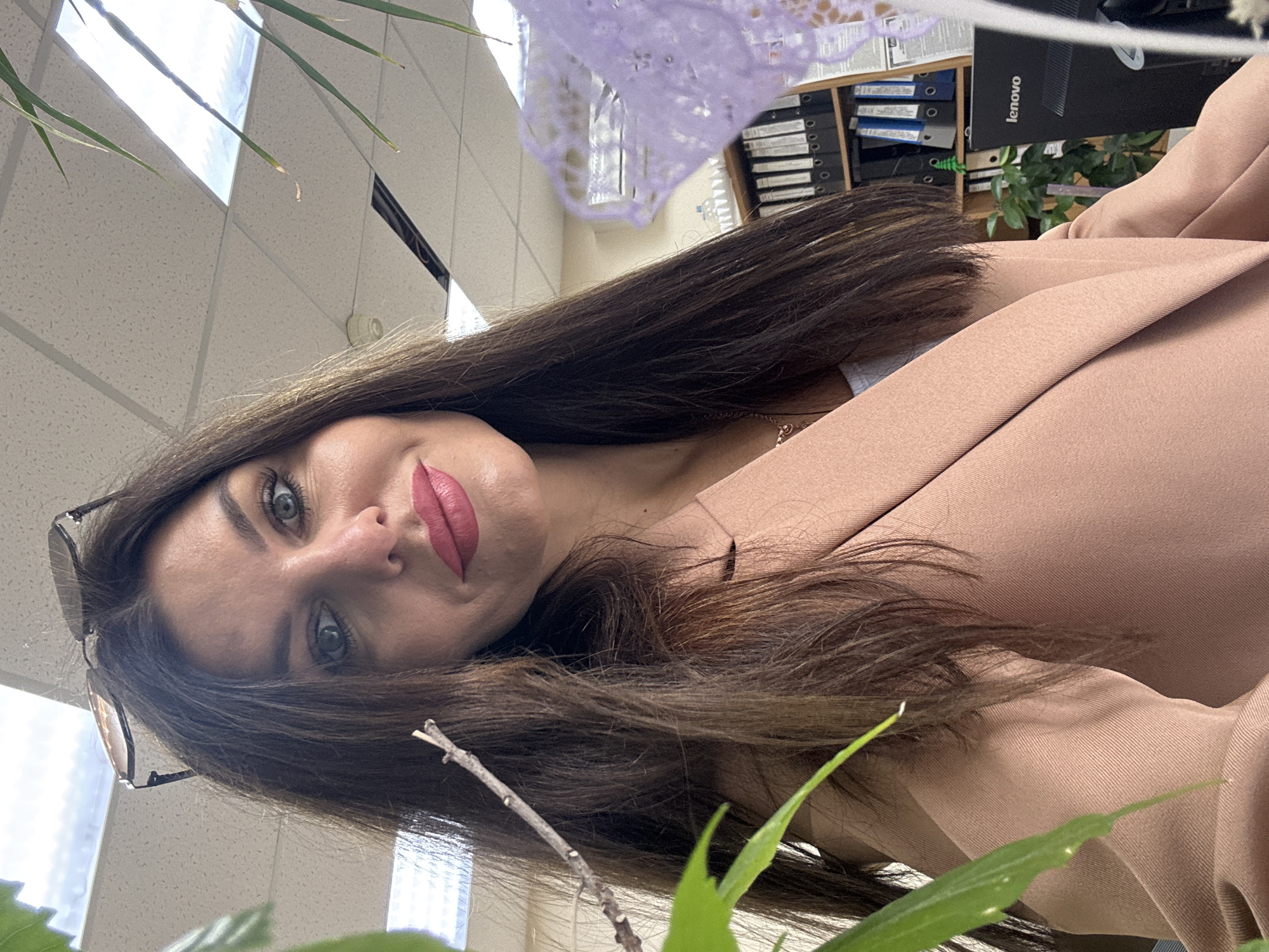 The image is a close-up, selfie-style shot of a woman in an office environment. She has long, dark brown hair, blue eyes, and is wearing pink lipstick. Her head is tilted slightly, and she is looking towards the camera. She is wearing a light pink blazer over a white top and a necklace. On top of her head, she has a pair of sunglasses. In the foreground, to the left of the frame, a green plant leaf is visible. In the background, there are office supplies, including binders on shelves, a computer monitor with the "Lenovo" logo, and a decorative purple item, possibly lace. The ceiling appears to be a standard office suspended ceiling with fluorescent lighting. The overall scene suggests an indoor office setting. The presence of a Lenovo monitor and the general office decor do not provide any specific indicators to pinpoint the exact location in the city of Chaikovsky, Russia.
