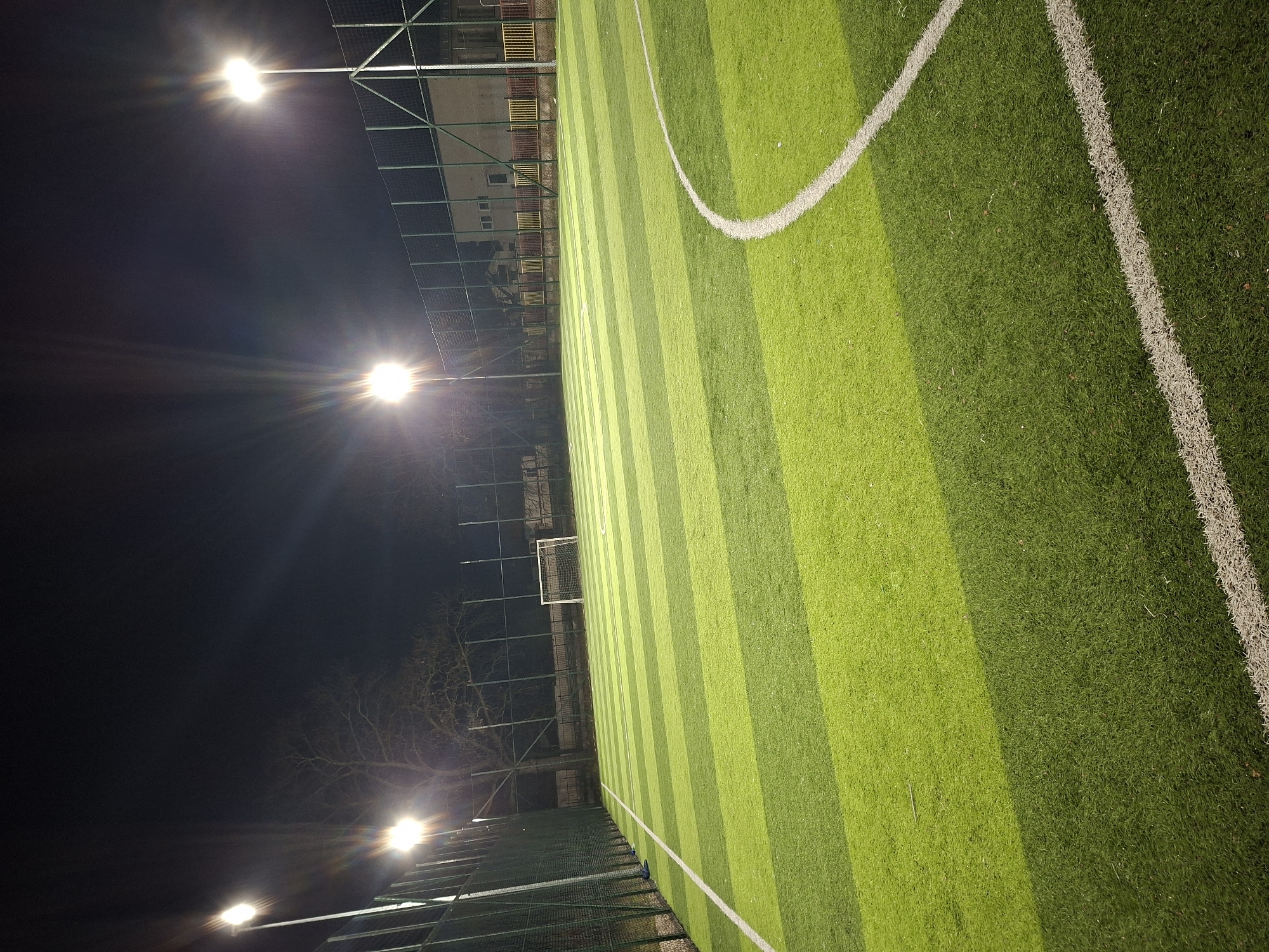 This image depicts an artificial turf soccer field at night, located in Zhitnitsa, Bulgaria. The field features alternating stripes of light and dark green synthetic grass, marked with distinct white lines including a straight sideline along the left edge and a curved arc in the foreground on the right. Four tall light poles, each equipped with bright floodlights, illuminate the scene from the left and right sides, casting significant lens flares. The sky above is dark, indicating nighttime conditions. A tall green mesh fence encloses the field, running along the left perimeter and continuing into the background. A white soccer goal with a net is visible at the far end of the field, centrally positioned. Behind the goal and fence, a light-colored building with windows and a door is present, further separated by a lower fence featuring yellow and red vertical bars. Bare trees are discernible behind the structures in the background. No individuals are present on the field or within the visible area, and no active play is observed. A small, dark blue object, possibly a shoe or bag, rests near the left sideline against the fence.