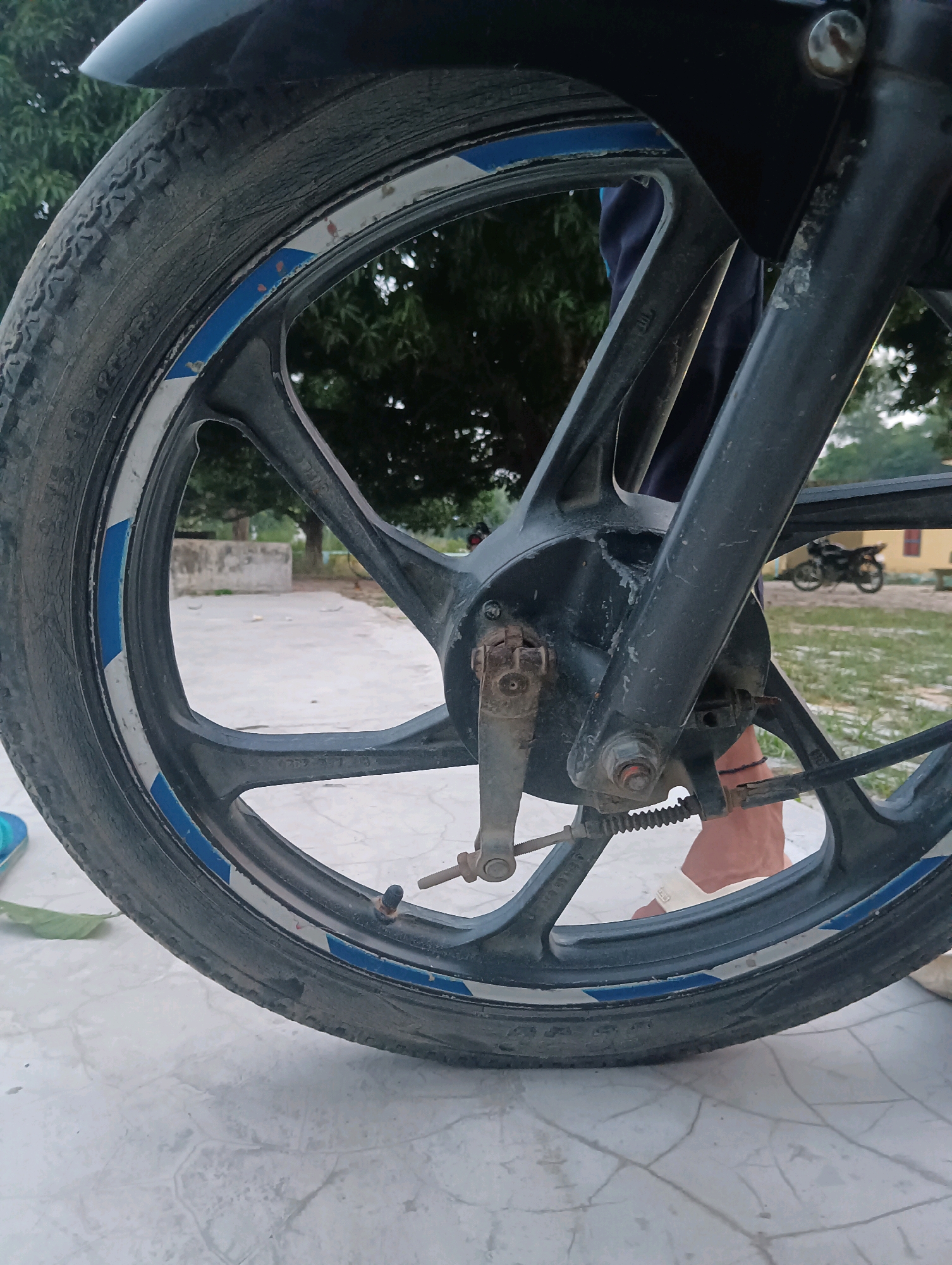 The image shows a close-up of a motorcycle's front wheel, seen from the left side. The wheel is black with blue and white decals on the rim. The tire is worn. The motorcycle is parked on a paved surface that appears to be outdoors. In the background, there are trees, a low concrete structure, and a distant building with a yellow wall. Another motorcycle is parked further in the background. A person's legs are visible behind the front wheel, with their feet wearing light-colored sandals. The scene is likely in Tilkar, India, given the background elements suggestive of a rural or semi-urban environment typical of such locations. The lighting suggests it is daytime, possibly late afternoon.