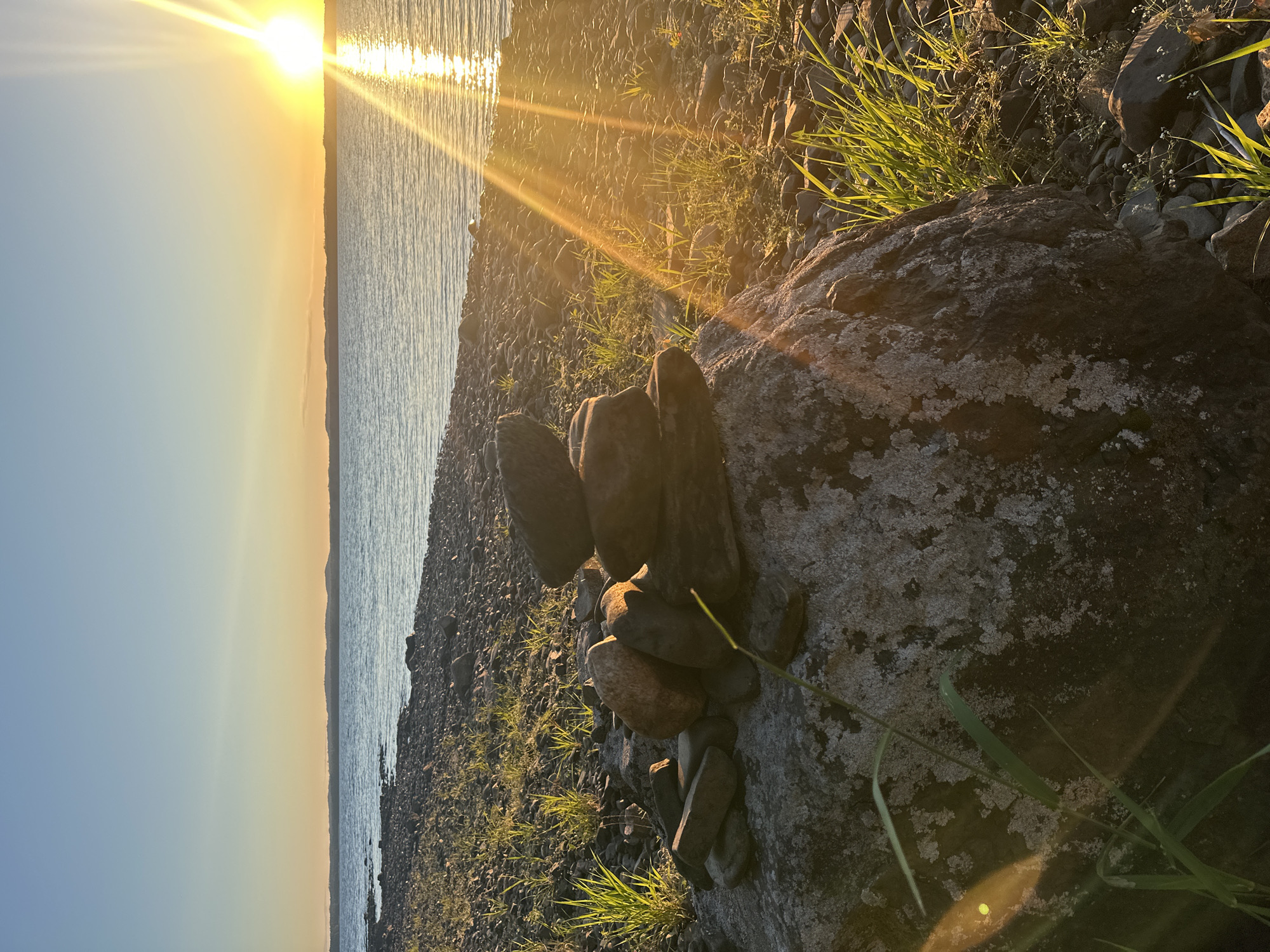 The image depicts a calm body of water under a clear sky at sunset. A rocky shoreline with scattered grass leads to the water's edge. In the foreground, a stack of balanced rocks, known as a cairn, sits on top of a larger boulder. The sun is low on the horizon, casting a warm, golden light and creating a shimmering reflection on the water. The scene appears to be a serene natural landscape, possibly located in the vicinity of Soanlakhti, Russia, known for its natural beauty and waterfront areas.