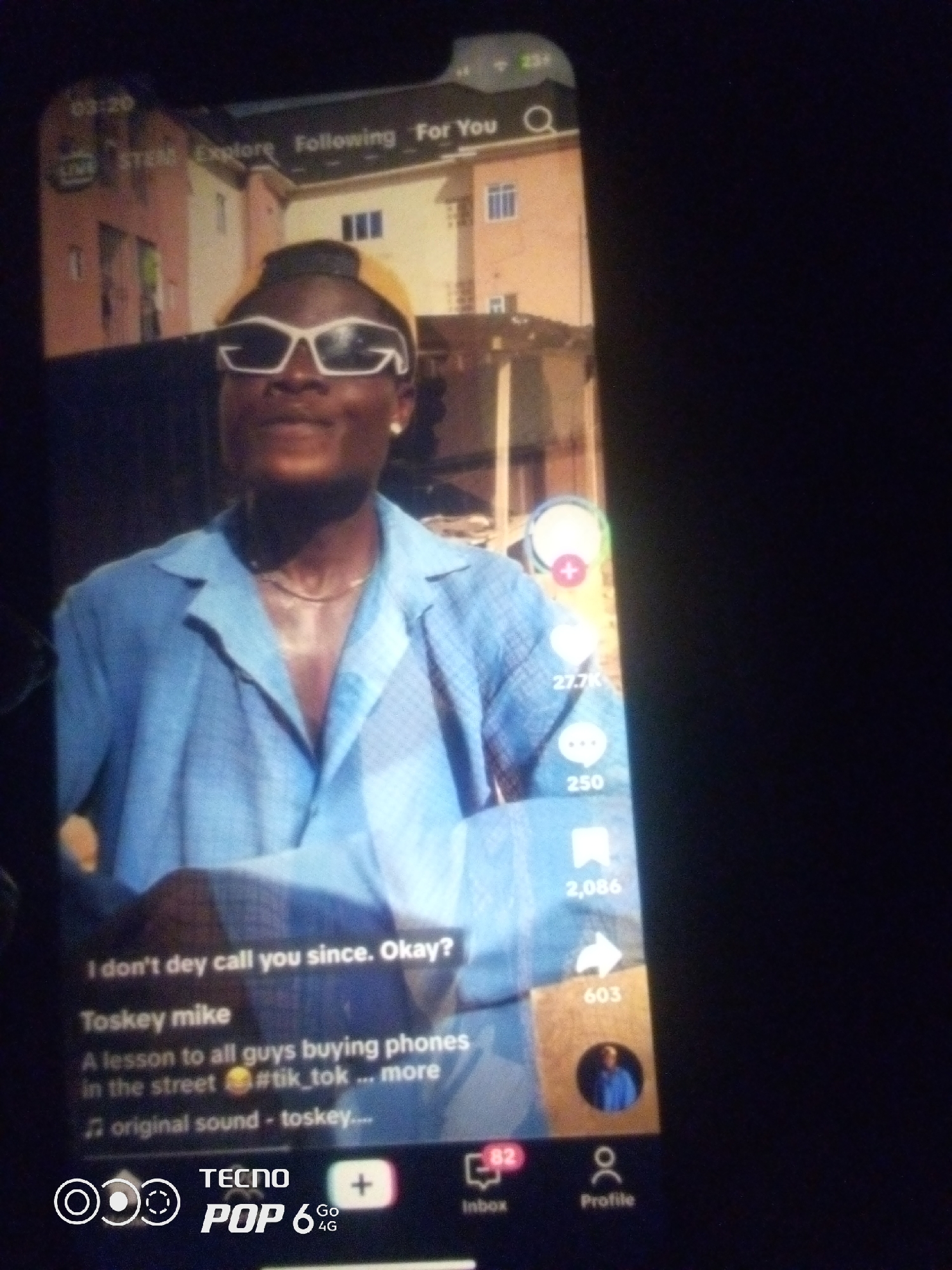 The image shows a screenshot of a TikTok video. A young man wearing a blue button-up shirt and sunglasses is the main subject. The background appears to be an urban environment with buildings. Text overlays on the video include a caption, username, and engagement metrics. The bottom of the image shows a phone's interface, specifically the TECNO POP 6 4G branding. There is no indication of the location being Howayi, Nigeria.