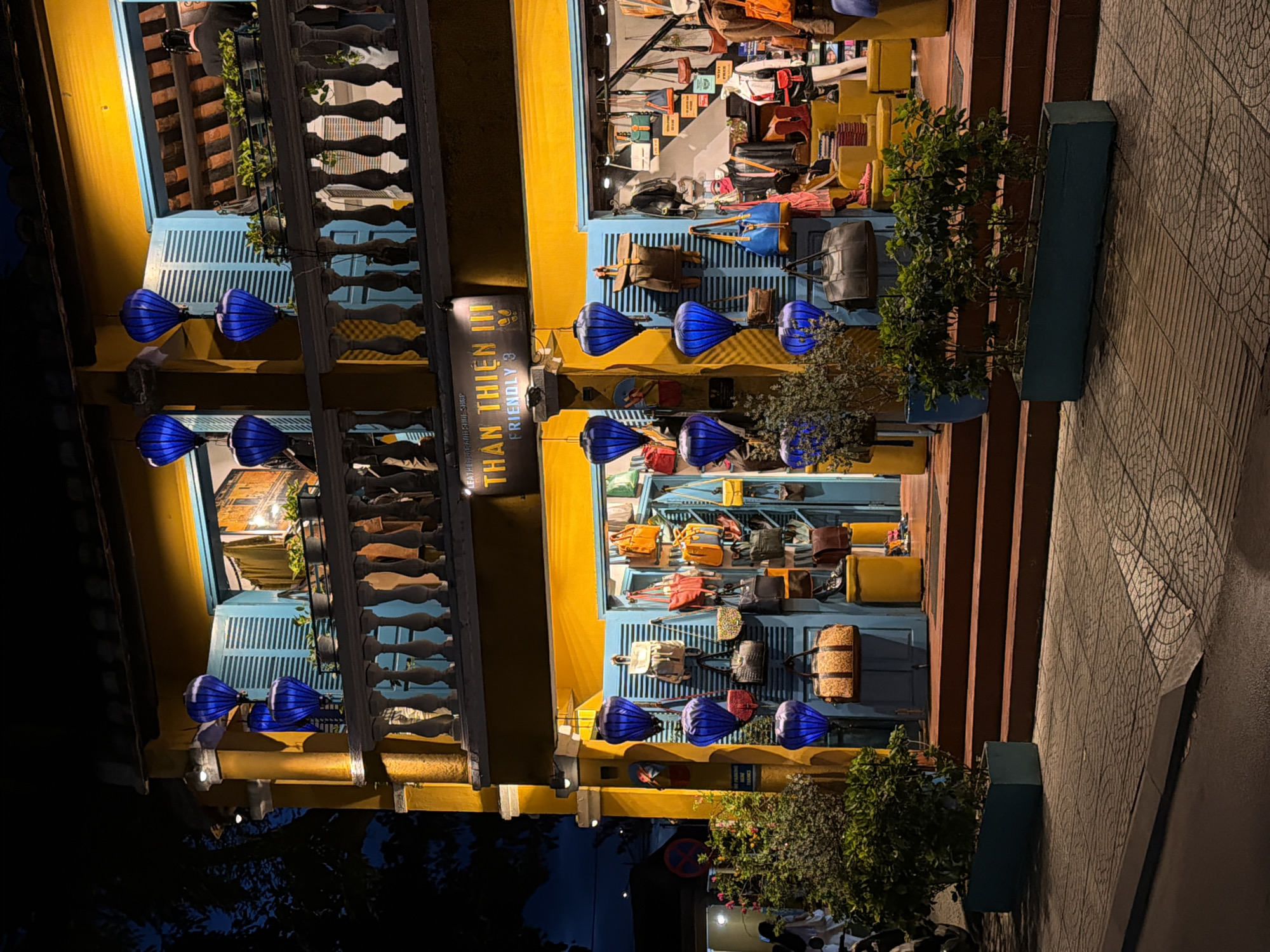 The image depicts a street-level view of a brightly lit shop with a yellow facade in Hoi An, Vietnam. The shop appears to sell bags, as many are displayed both inside the storefront and hanging from the exterior. Blue shutters are visible behind some of the bags. Hanging from a railing, several large, blue, bell-shaped lanterns illuminate the scene, suggesting it is evening or nighttime. A sign with Vietnamese text and "FRIENDLY 3" is also visible. The building has multiple stories, with balconies and railings. A tiled sidewalk runs in front of the shop, and stairs leading up to the entrance are visible on the right side of the image. Green plants in pots are placed near the entrance and along the sidewalk. The overall impression is a vibrant and inviting street scene at dusk or night.