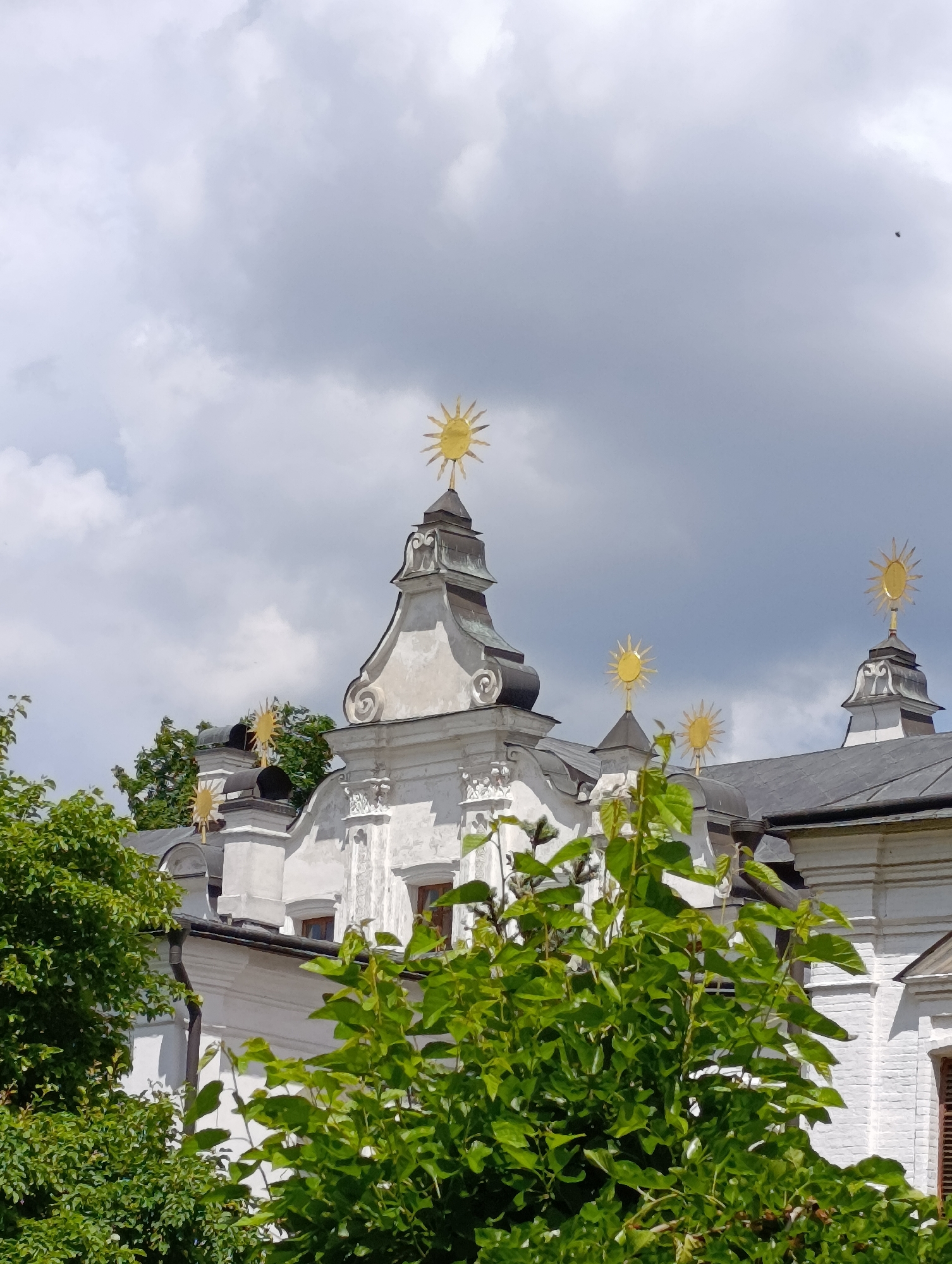This is a medium-shot, eye-level photograph of the rooftops of a white building, likely a church or monastery, in Kyiv, Ukraine. The architecture features multiple ornate domes topped with golden sun symbols. Lush green trees in the foreground partially obscure the view of the building. The sky is overcast with dramatic, textured clouds, suggesting a partly cloudy or stormy day. The lighting indicates it is daytime, possibly late morning or afternoon, given the diffused light. No people are visible, and there is no visible text in the image. The overall impression is one of historical architecture set against a dynamic natural backdrop.