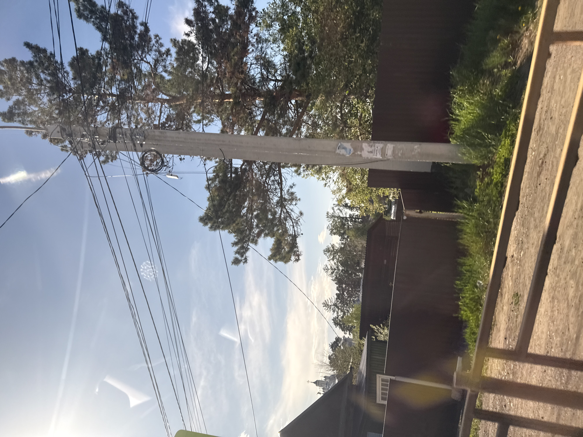 The image shows a view in Pivovarikha, Russia, featuring a tall concrete utility pole with numerous power lines extending across the sky. The pole is situated alongside a fence, with vegetation growing at its base. Beyond the fence are residential buildings and trees. In the distance, a church dome is visible against the blue sky with light clouds. The image is taken from a low angle, looking upwards towards the sky and the utility pole. There are no people visible in the image.
