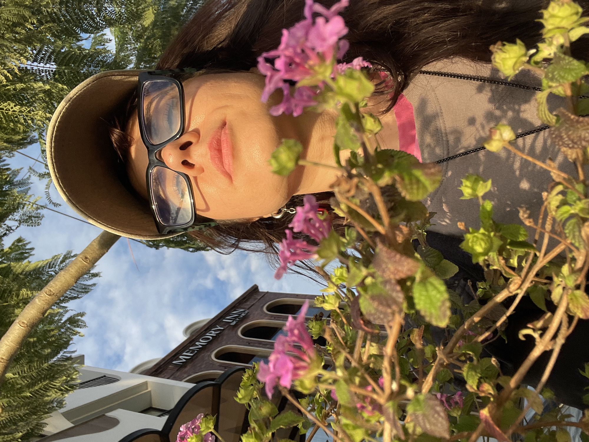 A low-angle, medium shot captures a woman wearing a baseball cap and glasses, her face tilted upwards towards the sky. She is partially obscured by the branches and pink flowers of a plant in the foreground. Behind her, a building with the words "MEMORY INN SKY VIEW" is visible, suggesting a location in Da Lat, Vietnam. The sky is blue with scattered clouds, and sunlight filters through the leaves of a tree overhead, casting shadows on the scene. The woman's expression is relaxed, and her eyes are closed, hinting at a moment of peaceful contemplation.