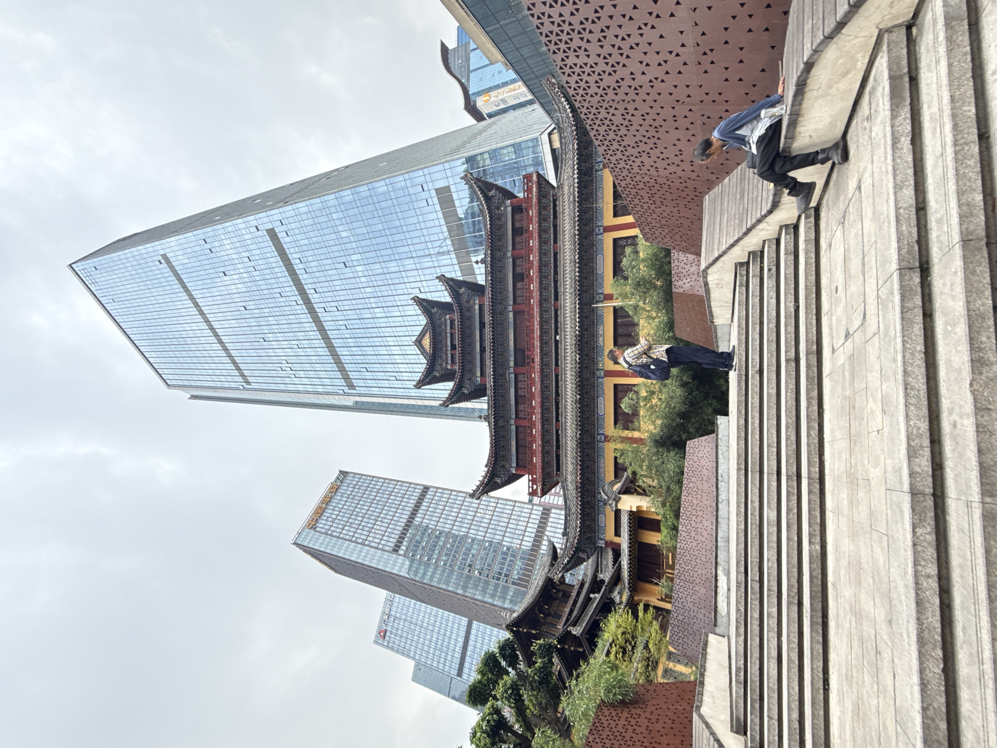 The image depicts a scene in the Yuzhong District of Chongqing, China. A traditional Chinese pagoda-style building is juxtaposed against modern high-rise buildings covered in glass. The architectural contrast highlights the blend of old and new within the city.
Two people are visible on a set of concrete steps in the foreground, one appearing to walk upwards. The scene is shot from a low angle, emphasizing the height of the surrounding structures. The sky is overcast, suggesting a cloudy day.