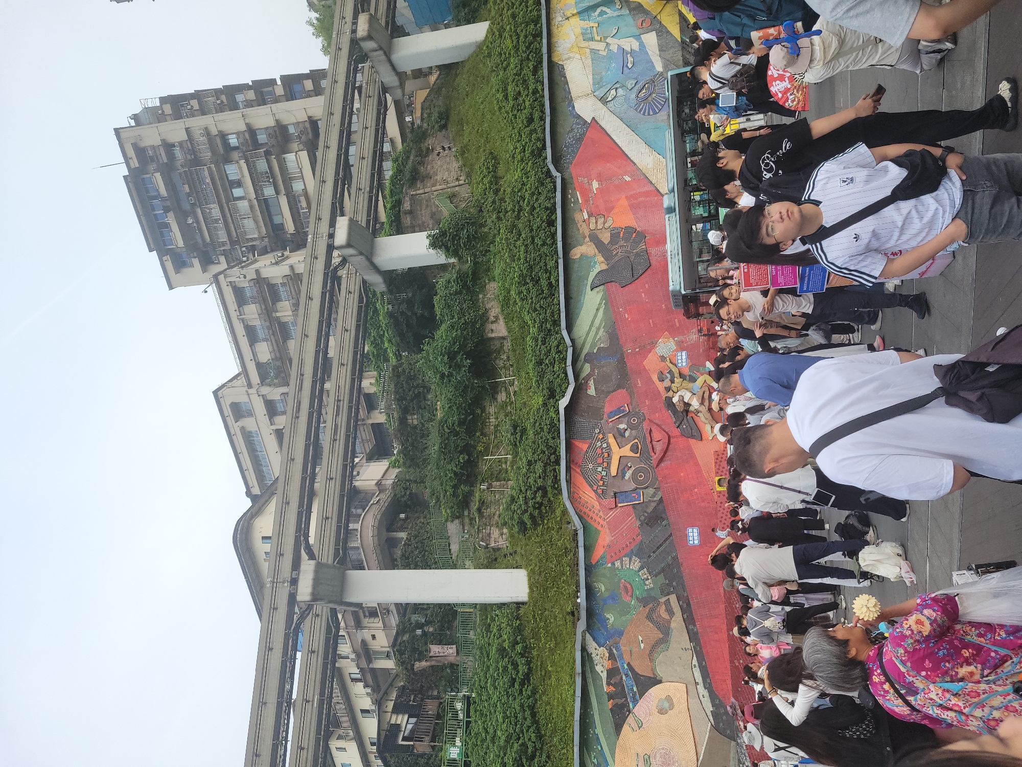 The image captures a crowded scene in Yuzhong District, China. A diverse group of people are standing along an expansive, colorful mural. The mural depicts a variety of stylized figures and geometric shapes. To the left of the mural, a monorail track runs above a hillside covered in dense greenery. In the background, tall residential buildings rise into the sky. The weather appears overcast.