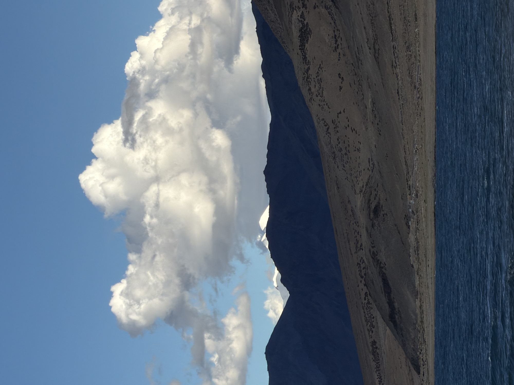 A vertical, eye-level, medium shot captures a serene landscape with a body of dark blue water in the foreground. The water's surface is rippled, suggesting a gentle breeze. Beyond the water, a barren, brown hillside slopes upwards, dotted with sparse vegetation. A range of dark, silhouetted mountains forms the mid-ground. In the distance, snow-capped peaks are visible between the mountain range and the sky. The sky is a vibrant blue, adorned with large, fluffy white clouds that cast shadows on the mountains below. The scene is devoid of any human presence or structures.