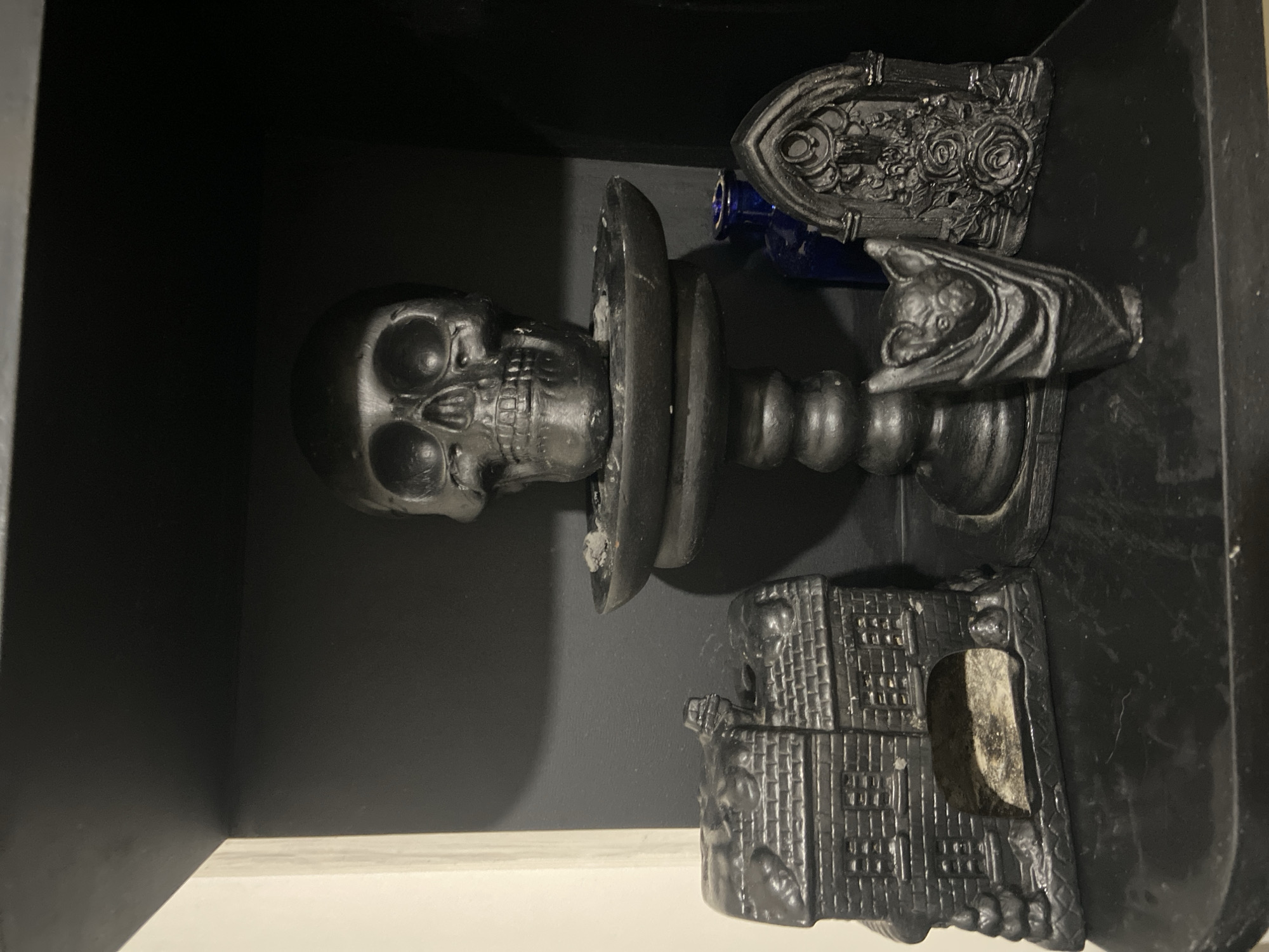 This media file presents a still life arrangement of dark-themed decorative objects displayed within a dark-colored shelf or cubby. The scene is indoors, characterized by uniformly dark walls and a lighter, possibly off-white, surface at the bottom supporting some of the items.
The central subject is a matte black human skull, facing forward, prominently displayed on an ornate, black candlestick-like stand. The skull is stylized but detailed, resting on a flat, circular platform that forms part of the stand. Attached to the side of this candlestick, a small, black bat figure is visible, perched with its head facing right.
To the right of the skull and behind the candlestick, a dark blue glass bottle or vial is partially obscured by a rectangular, black ornamental plaque. This plaque features intricate raised designs of roses and leaves, consistent with a gothic aesthetic.
In the lower right corner, resting on the lighter shelf, is another black object resembling a miniature, weathered building or sarcophagus. It exhibits textured details that suggest stone or brickwork and has a rectangular opening revealing a lighter, possibly off-white, interior texture.
There is no activity or event taking place; the image captures a static display. The overall tone is dark and somewhat macabre, consistent with gothic decor. The lighting highlights the textures of the objects against the dark background. No visible text is present in the image.
