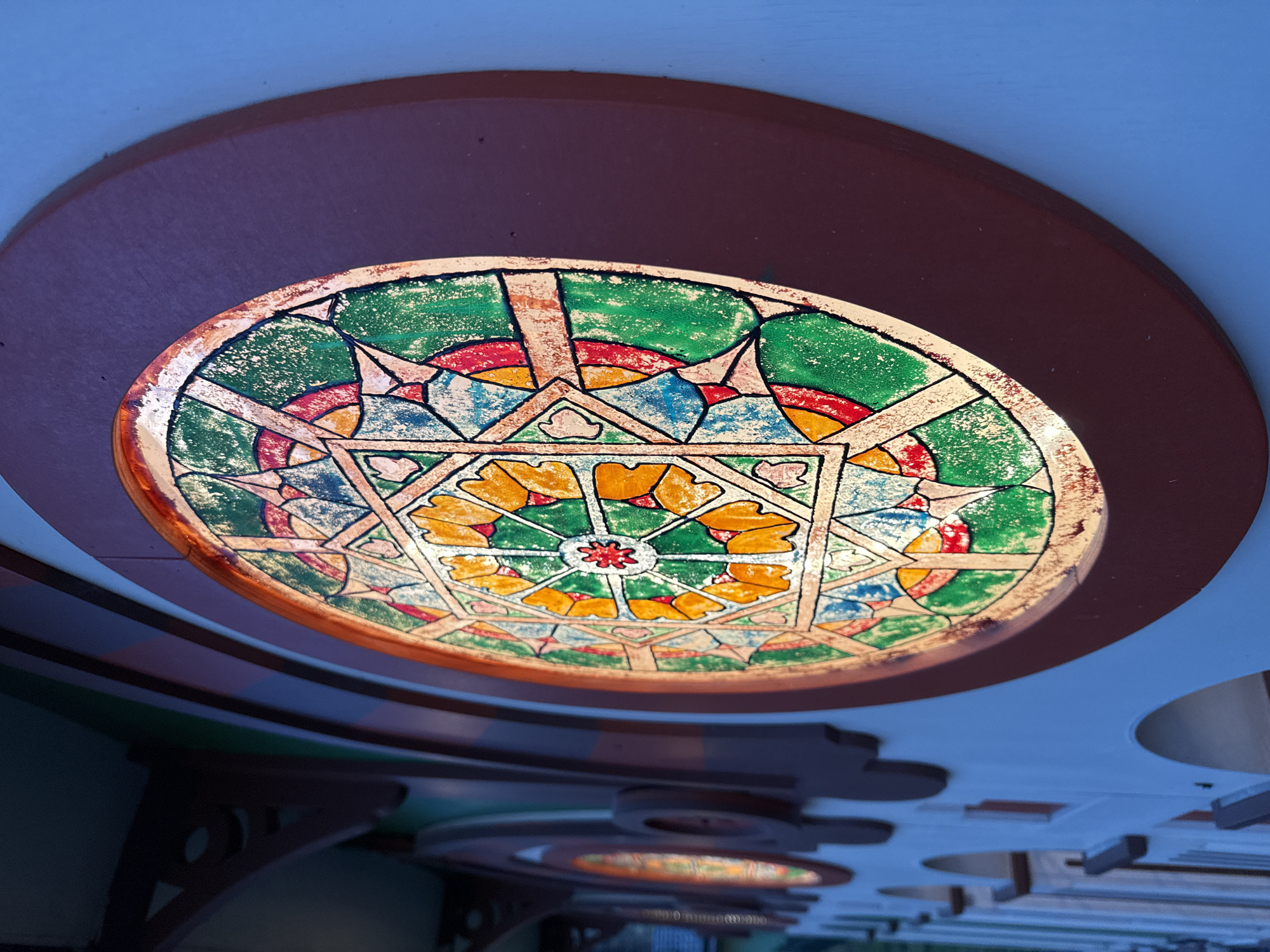 The image shows a stained glass ceiling fixture in an interior space. The fixture is round with a geometric pattern in shades of green, red, blue, yellow, and white. A brown ring surrounds the fixture, and the surrounding ceiling is pale blue. In the background, there are glimpses of additional fixtures and architectural details, suggesting a public building. While I cannot definitively identify the exact location as Perm, Russia, the architectural style suggests it may be within a historical building somewhere in Russia.