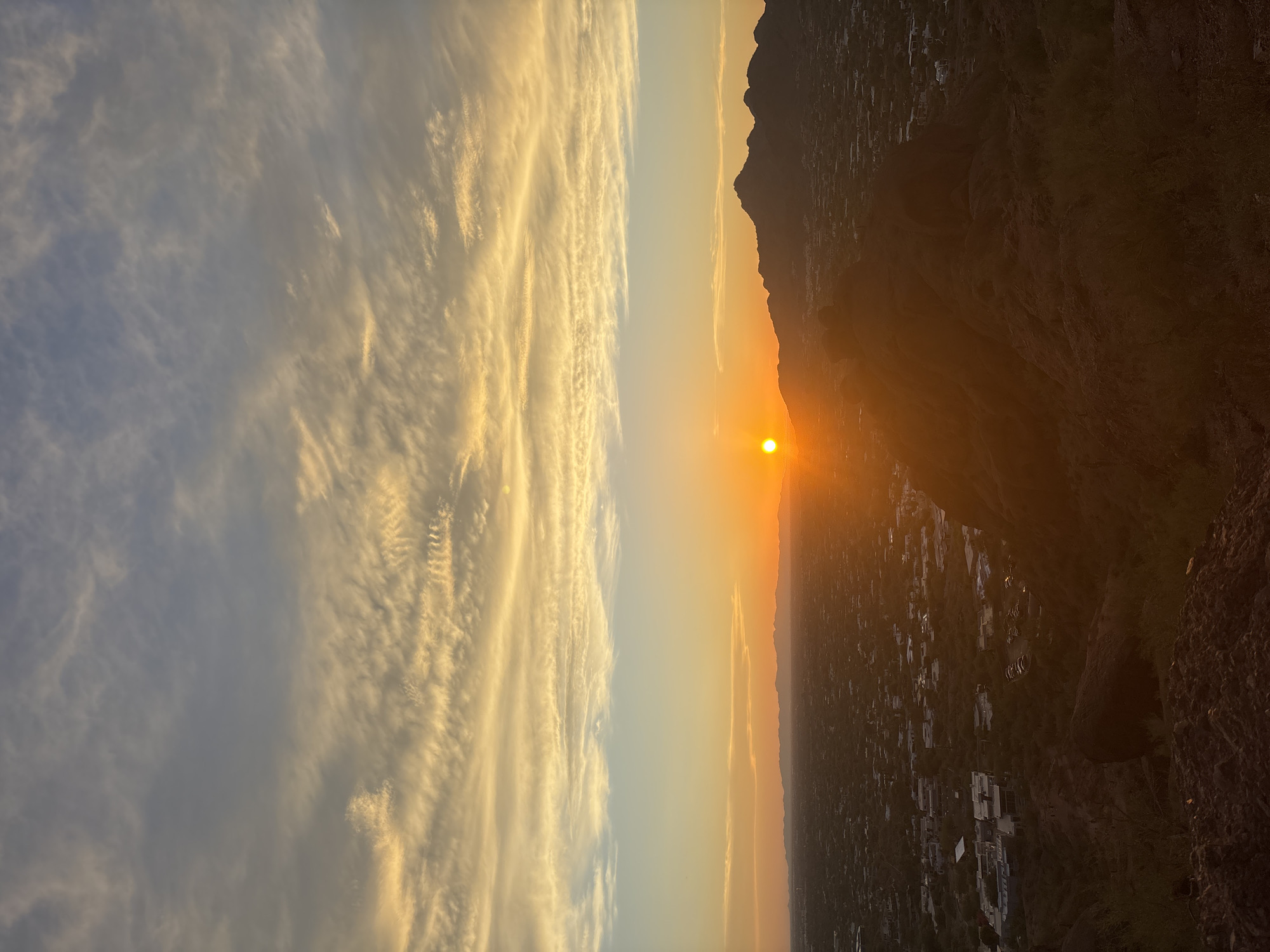 The image captures a sunset viewed from a high vantage point, likely a mountain or hill, overlooking the city of Phoenix, Arizona, USA. The sun is setting on the horizon, casting a warm orange glow across the sky and illuminating the clouds with streaks of yellow and white. In the foreground, the dark silhouette of a rocky terrain with sparse vegetation is visible. Below, the city sprawls out, a tapestry of buildings and structures, with distant mountain ranges forming the backdrop. The scene is characterized by the dramatic interplay of light and shadow, with the setting sun creating a vibrant and atmospheric panorama.