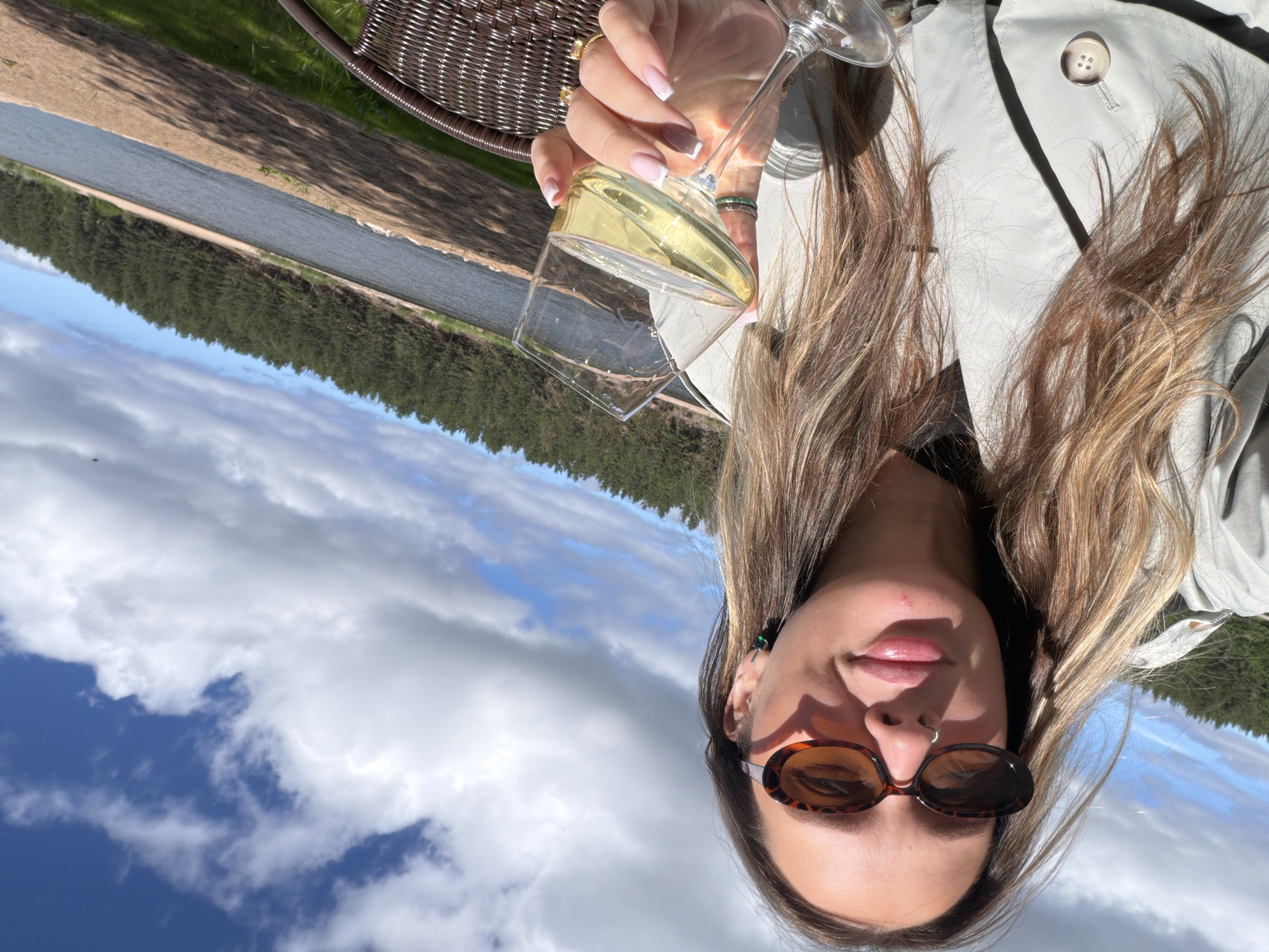 This media file features a young woman holding a glass of white wine, with a scenic natural landscape in the background. The woman is wearing sunglasses and a light-colored trench coat. Her hair is long and blonde, framing her face. She is smiling faintly. The setting appears to be outdoors, with a body of water, a line of trees, and a cloudy blue sky visible. The orientation of the image is sideways. The time of day is likely daytime, given the bright sunlight casting shadows. The weather seems pleasant, with a mix of sun and clouds. The scene evokes a sense of relaxation and enjoyment, possibly a vacation or a leisurely outing. The specific location is identified as Solov'evo, Russia. There is no visible text in the image.