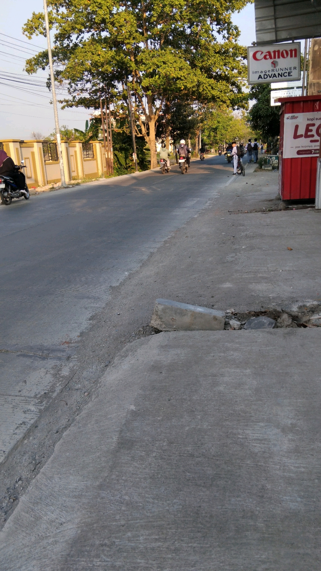 The image depicts a street scene in Tegal, Indonesia, during daylight. A paved road runs diagonally from the lower left to the upper right. To the left of the road, a person is riding a black motorcycle. Further down the road, several other individuals on motorcycles are visible. A white building with a sign that reads "Canon" is situated on the right side of the road, with a red kiosk next to it displaying text that appears to be in Indonesian. The foreground features a cracked concrete sidewalk with debris. Lush green trees line the right side of the road, casting shadows. The sky is clear and bright, suggesting fair weather.