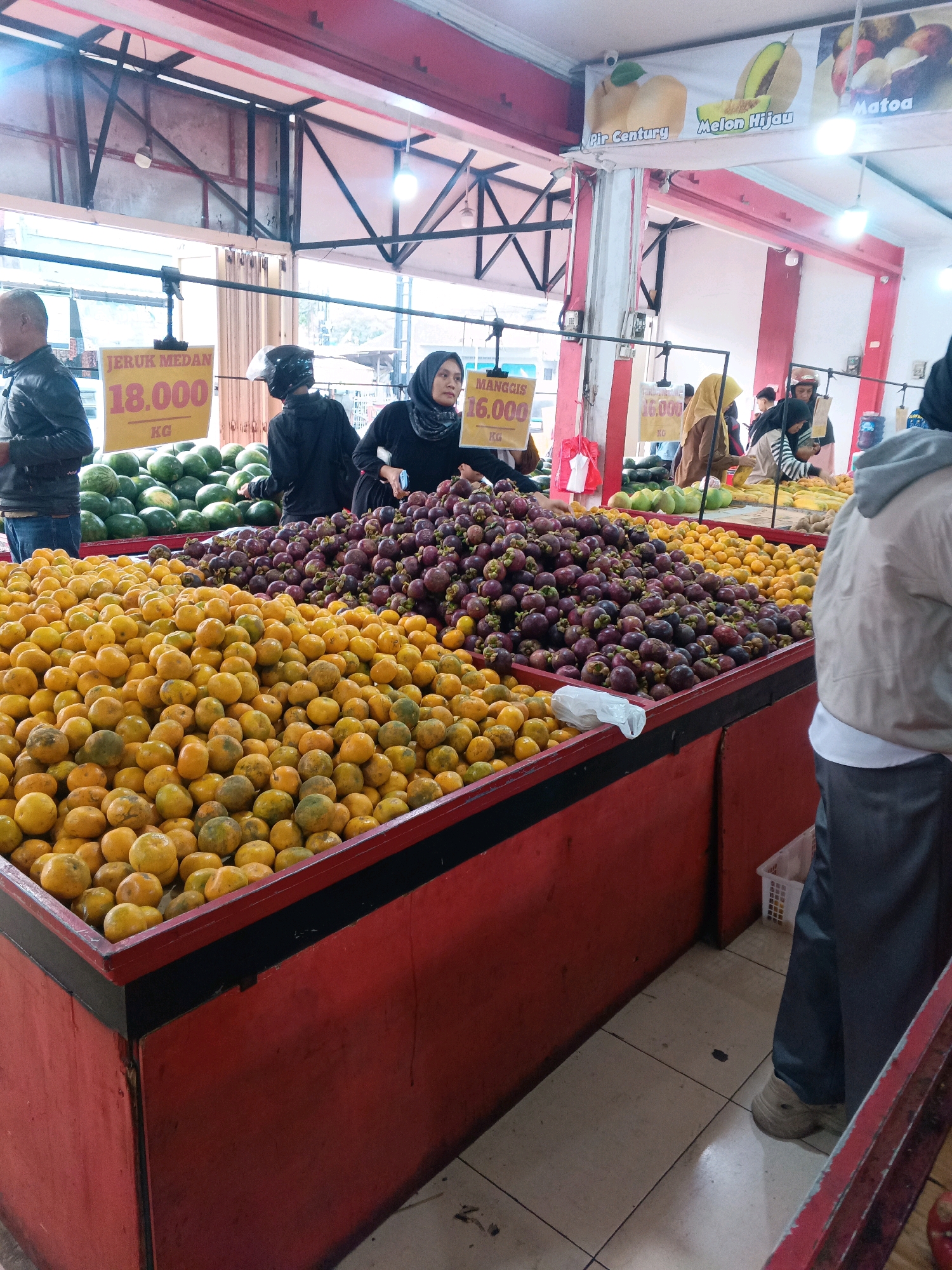 In an indoor fruit market, likely located in Garut, Indonesia, a variety of fruits are displayed in large red bins. The foreground is dominated by a bin overflowing with bright orange tangerines. Behind the tangerines, a bin is filled with dark purple mangosteens. Further back, a large bin holds numerous green watermelons. Signs indicate prices: one sign reads "JERUK MEDAN 18.000 KG" and another, partially obscured, reads "MANGGIS 16.000 KG". A banner above displays images of fruits, including "Pir Century", "Melon Hijau", and "Matoa". Several individuals are present. On the left, a man in a black jacket stands near the watermelon bin. In the center, a woman wearing a black hijab and black clothing is seen talking to another person. To the right, a person wearing a grey hoodie and dark pants stands facing away from the camera, observing the fruit. The market appears to be well-lit with artificial lighting. The flooring is tiled.