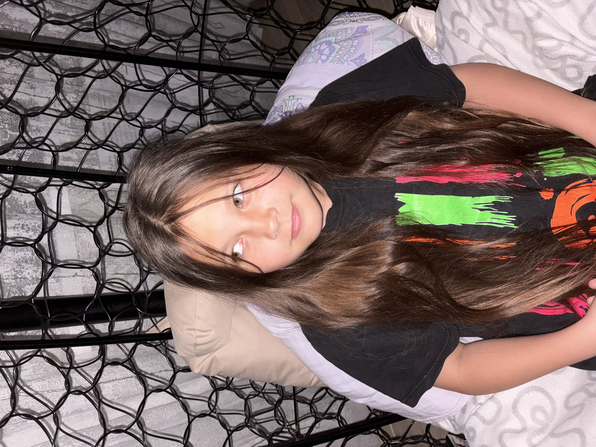 A young girl with long brown hair lies on a bed. She is wearing a black T-shirt with colorful graphics. The bedding includes a patterned pillow, a neutral-colored pillow, and a white sheet. A black metal bed frame surrounds the scene.