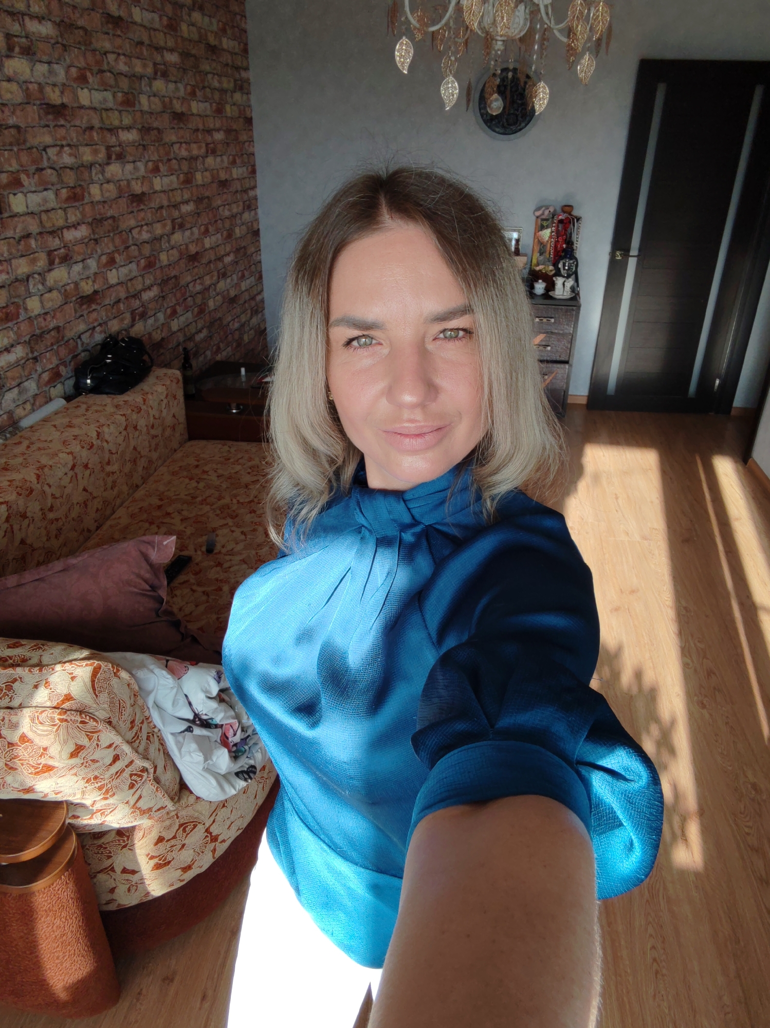 A woman takes a selfie indoors, looking directly at the camera with a subtle smile. She has light blonde/ash blonde hair, light green-brown eyes, and fair skin, and is wearing a long-sleeved teal blue top with a high neckline and pleated details. Her right forearm is visible in the lower right, holding the camera.
The setting is an indoor room, likely a living room or bedroom. The background features a distinctive brick-patterned accent wall on the left, adjacent to a plain gray wall. A sofa with an ornate floral pattern and a pink pillow occupies the left side of the room. On the right, a dark wooden dresser is topped with various decorative items and bottles, positioned next to a dark brown door with two vertical panels. A decorative chandelier with gold leaf-like ornaments hangs from the ceiling, along with a circular wall clock. The room has a light wooden floor.
Strong sunlight streams into the room, casting prominent, long shadows across the floor and furniture, indicating either morning or late afternoon. A black bag or helmet-like object rests on the sofa near the brick wall. No text is visible in the image.