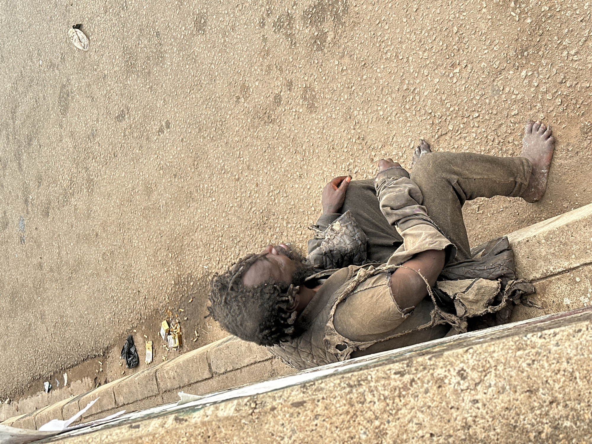 An overhead, high-angle image captures a single adult male sitting on a concrete curb next to a textured, light-colored wall or barrier. The individual, of apparent African descent, has dreadlocks and a beard, and his skin appears dark. He is dressed in tattered, earth-toned clothing, including a torn vest or jacket over a t-shirt, and long pants, all visibly soiled and worn. His bare feet, dirty and calloused, rest on the light brown, pebbled asphalt road surface. He is positioned with his knees bent, and his head is turned to his right, looking slightly upward. The area along the curb features discarded cigarette butts, yellow and white paper packaging scraps, a small black plastic bag, and some dark, possibly damp, patches on the pavement. A single light-colored leaf or piece of debris is visible further on the road surface in the upper right quadrant. The scene depicts an outdoor setting in an unknown city and country, with the individual in a state of rest or waiting amidst urban debris.